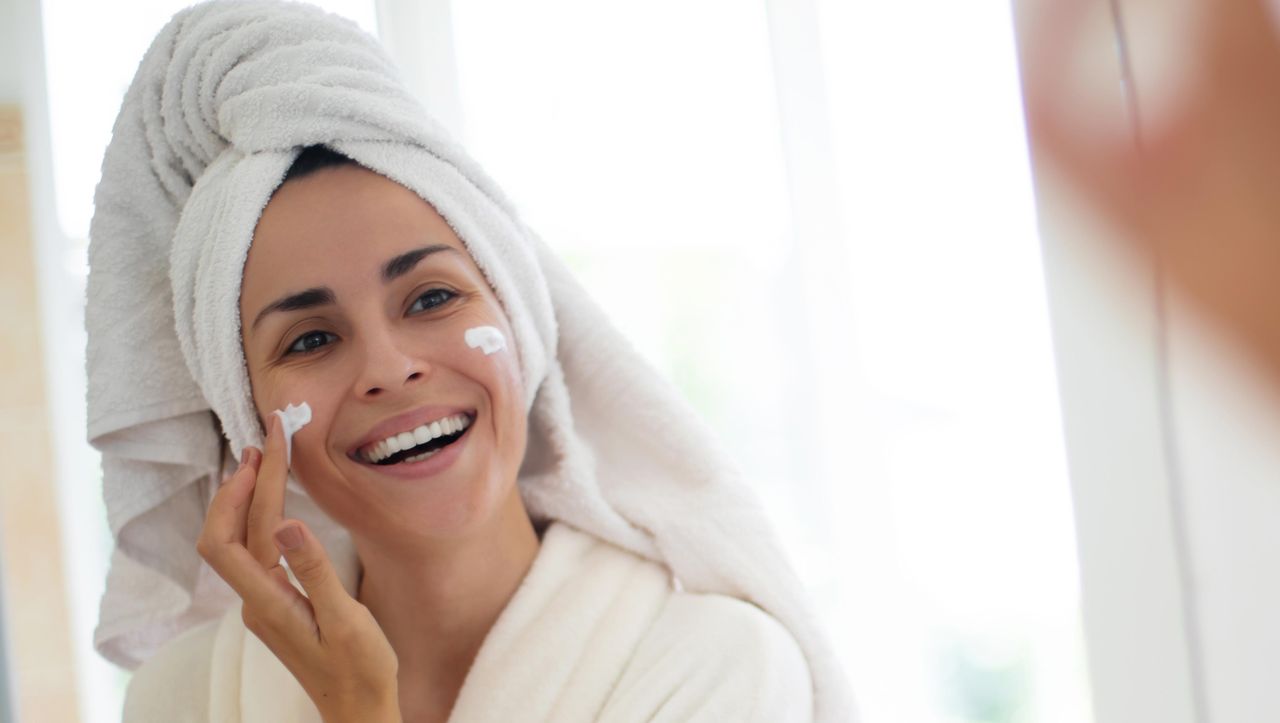 Perch_scegliere_una_crema_idratante_di_qualit_5lbAY8HNOn - Confidenze24 source:usmagazine - Perché scegliere una crema idratante di qualità - Una donna sorridente in accappatoio applica crema sul viso davanti a uno specchio del bagno.