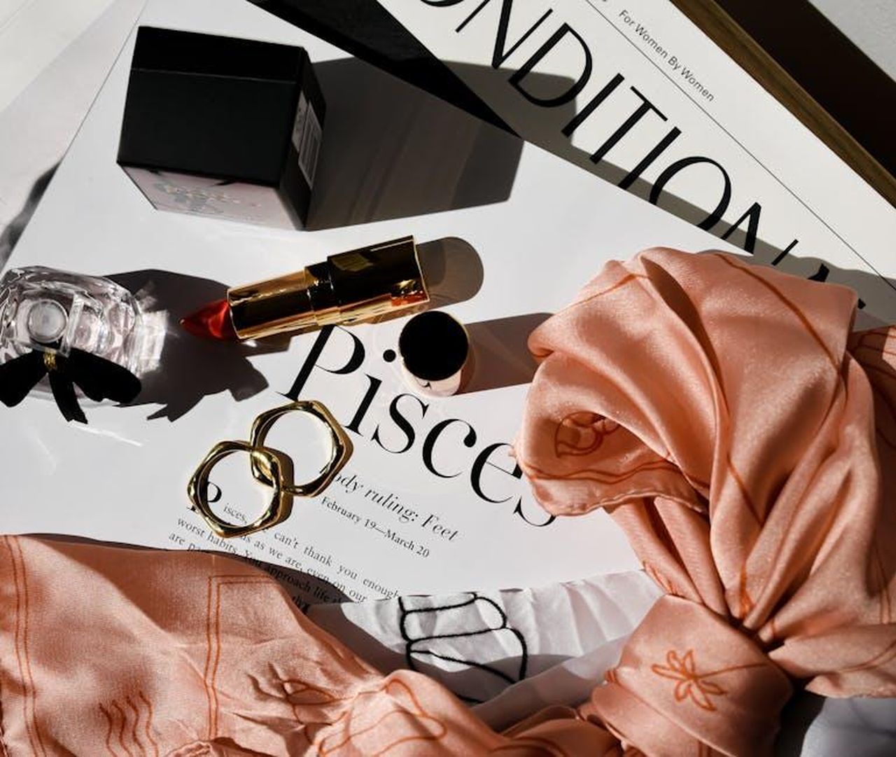 11_Pesci_generosit_nei_sentimenti_b6fxm3y4o7 - Confidenze24 Source: Harper Sunday - pexels - Chic flat lay featuring gold rings, red lipstick, perfume, and a stylish scarf over a fashionable magazine.