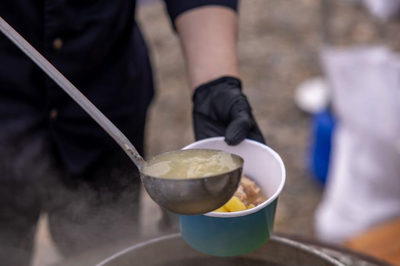 2018_Rescue_Dinner_UXxkIGsItN - Confidenze24 Source: Sergei Starostin - pexels - A person serves steaming hot soup from a pot into a bowl using a ladle outdoors.