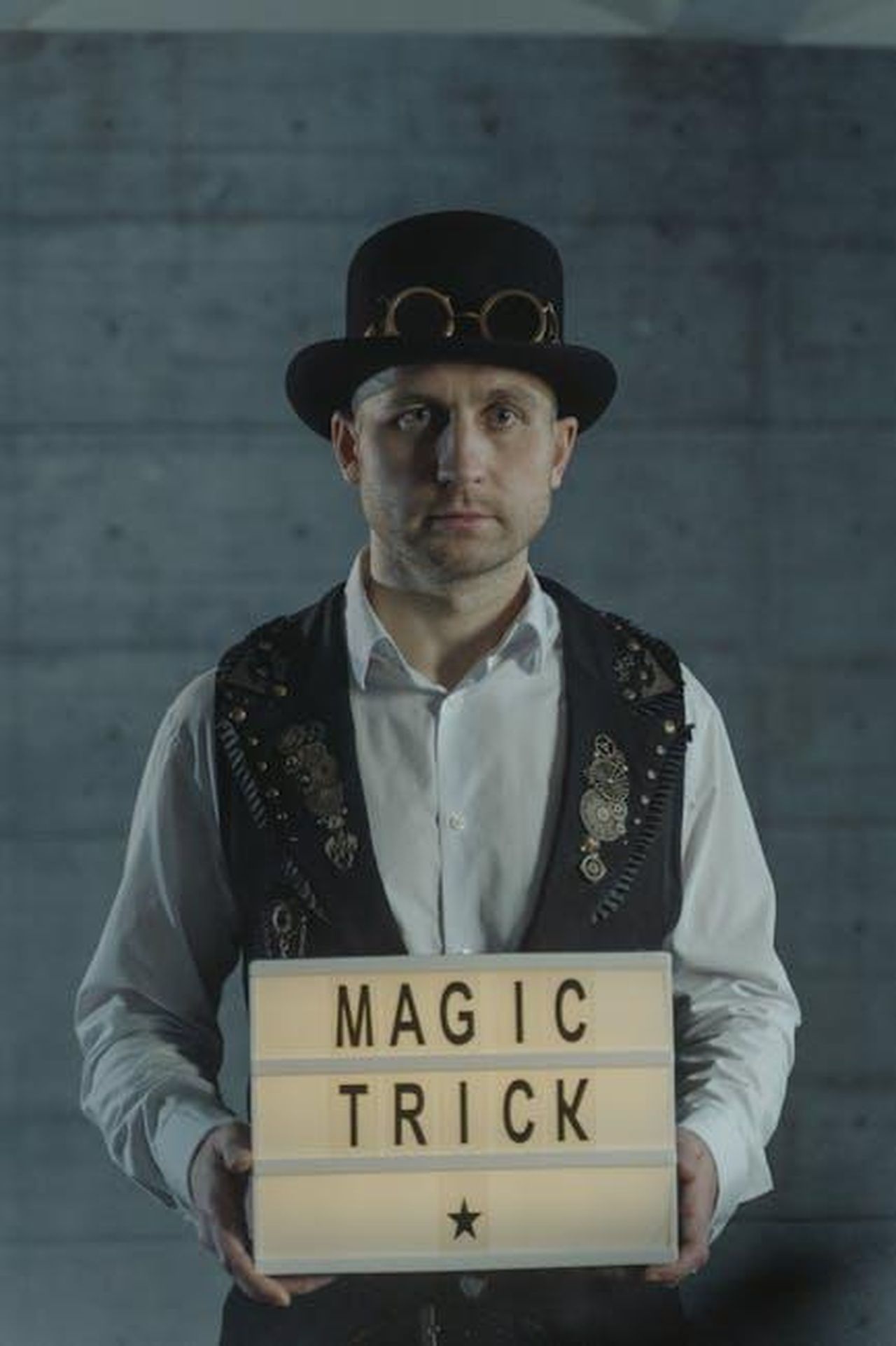 Buon_viaggio_JzC8sJOmew - Confidenze24 Source: Tima Miroshnichenko - pexels - A magician wearing a vintage hat holds a sign that reads 'Magic Trick' in an indoor setting.