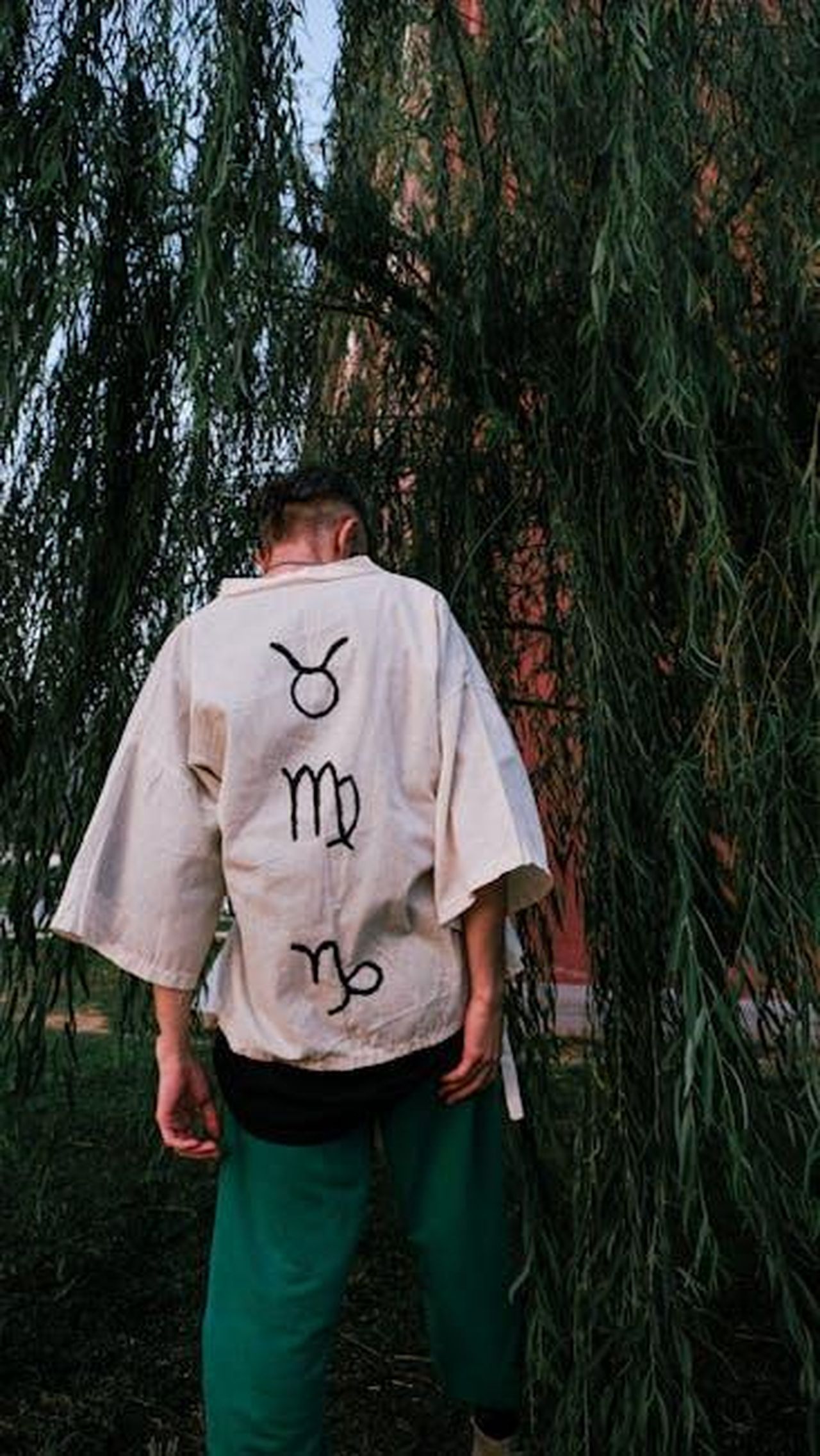 Capricorno_instabilit_e_confusione_aXr1O9vD9B - Confidenze24 Source: armağan başaran - pexels - A man stands outdoors wearing a shirt with zodiac symbols, surrounded by willow trees.