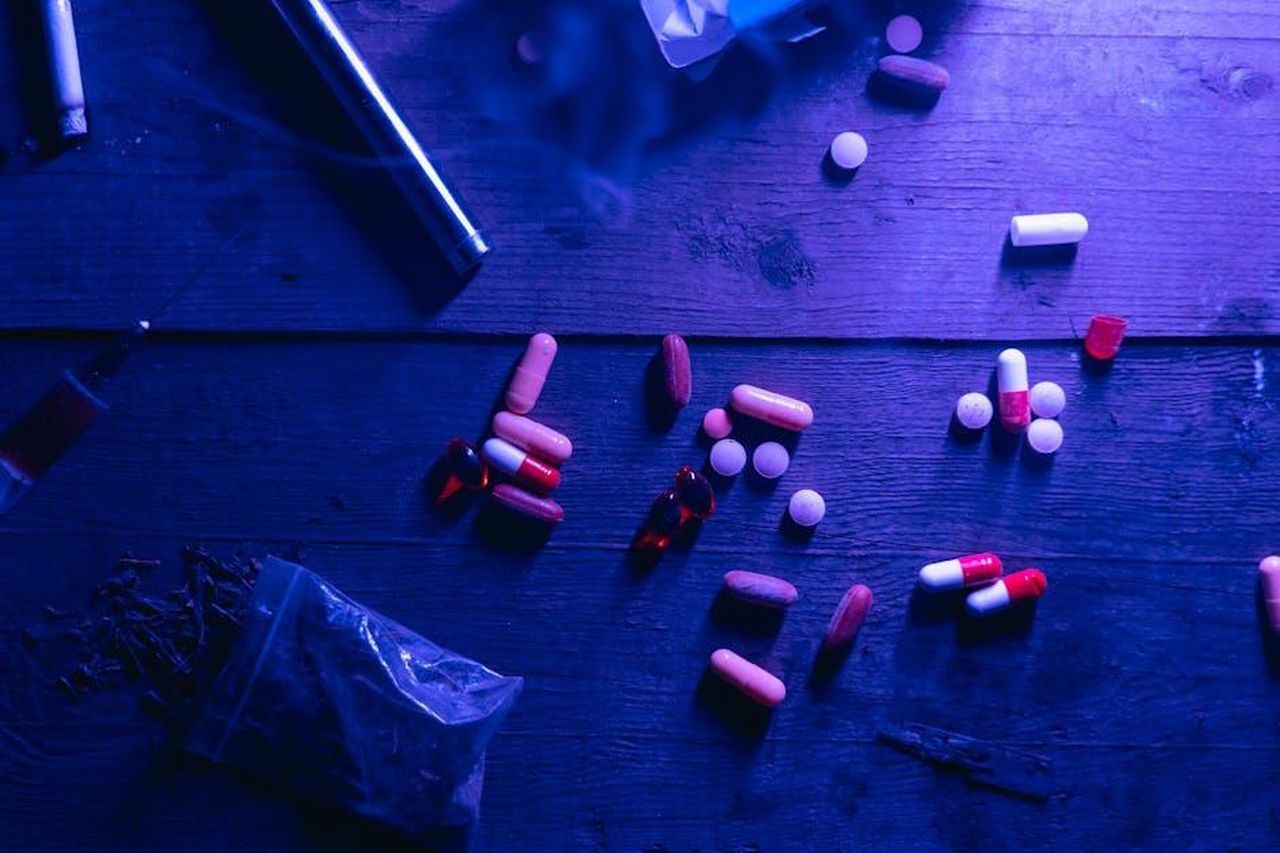 Causa_della_morte_e_problemi_di_salute_bucI9z9nmE - Confidenze24 Source: MART PRODUCTION - pexels - A flat lay of various drugs including pills, capsules, and a syringe on a wooden surface under moody blue lighting.