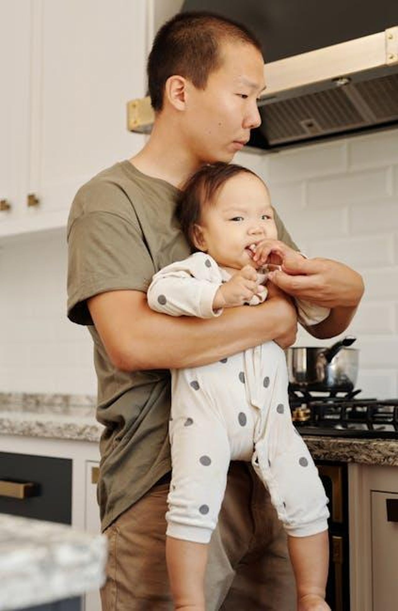 Christopher_Allen_Hackman_I4scG1rdRo - Confidenze24 Source: Vanessa Loring - pexels - Father lovingly holds baby in a modern kitchen, showcasing family bonding at home.
