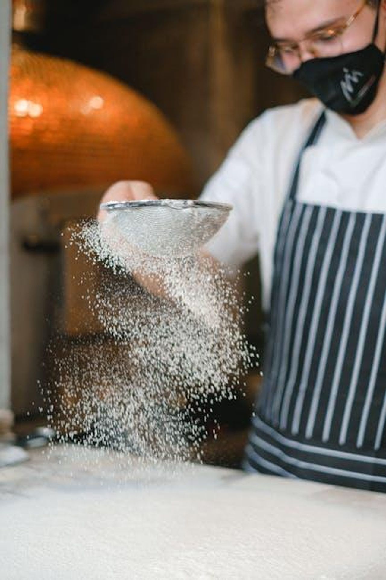 Come_preparare_i_breadcrumbs_allaglio_bcZ8agSL3c - Confidenze24 Source: Anna Shvets - pexels - A chef wearing an apron sifts flour onto a preparation surface in a professional kitchen.