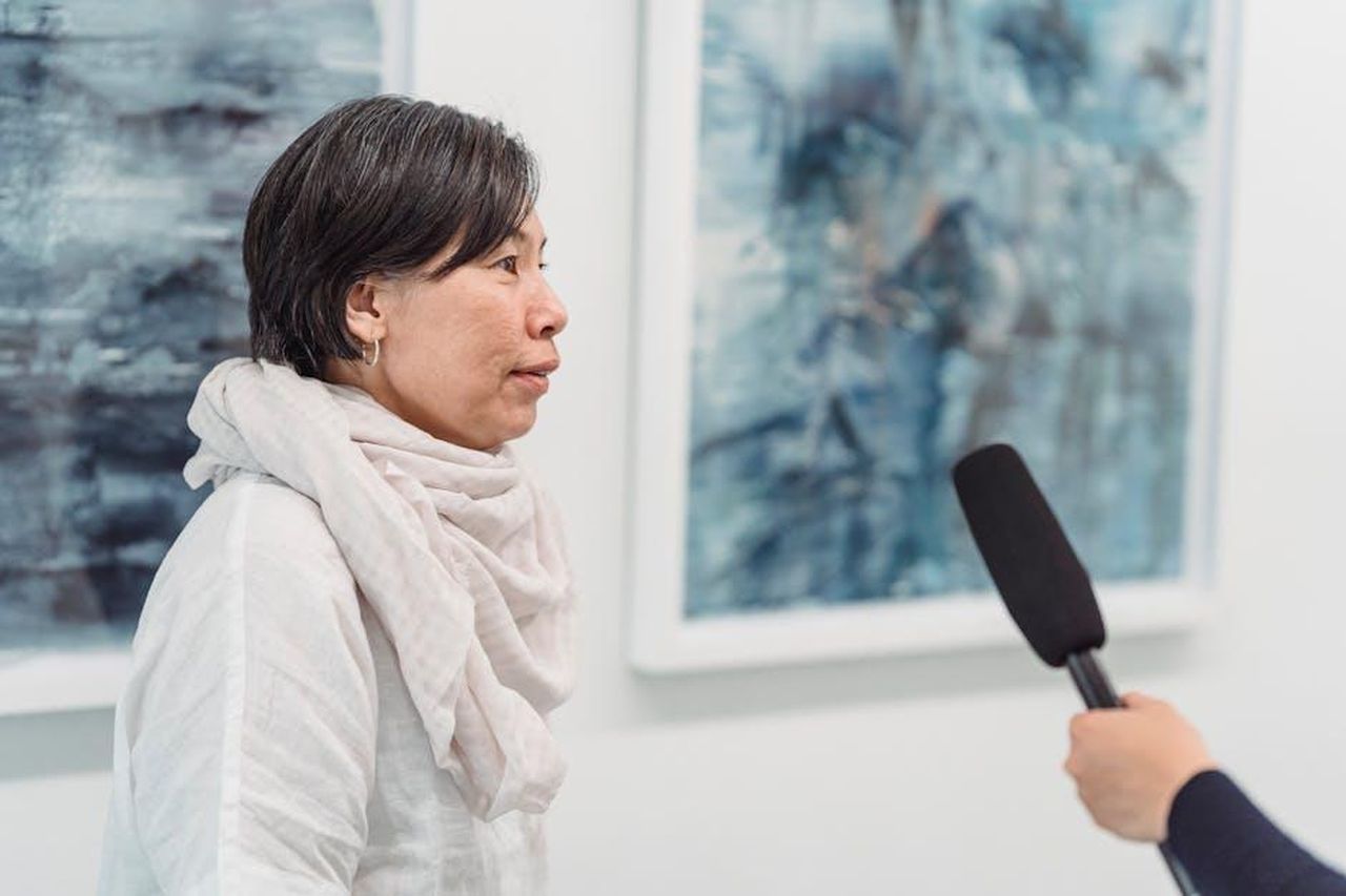 Critiche_allintervista_di_Panorama_NY6pggCJOJ - Confidenze24 Source: Greta Hoffman - pexels - A woman being interviewed in front of abstract paintings with a microphone.