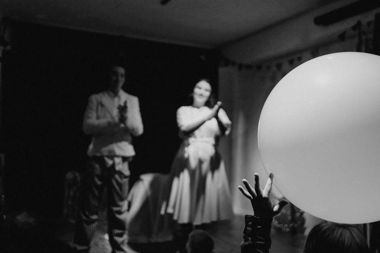 Dinamiche_tra_i_concorrenti_del_Grande_Fratello_YAnhqsvElj - Confidenze24 Source: Rina Mayer - pexels - Monochrome photo of performers on stage with a child's hand reaching for a balloon.