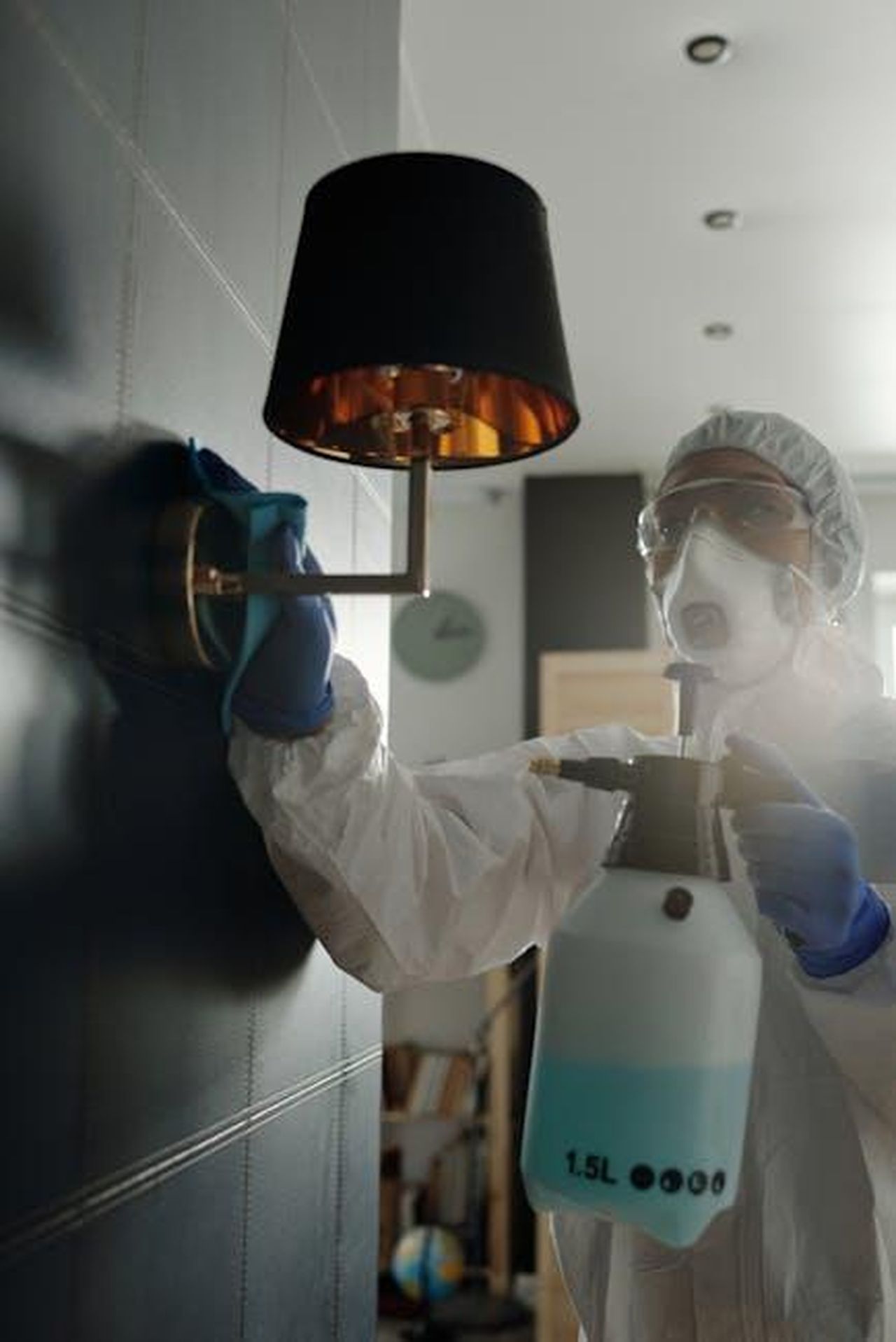 Due_esclusioni_in_pochi_giorni_tfqx36N7RW - Confidenze24 Source: Matilda Wormwood - pexels - Person in PPE sanitizing a wall-mounted lamp indoors with a disinfectant spray.