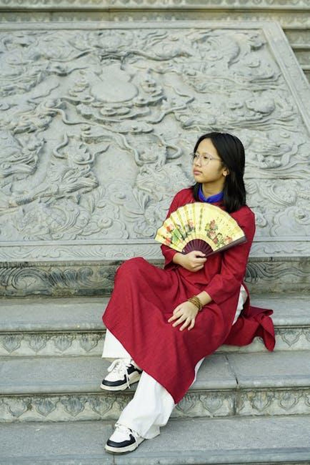 Fan_di_Upas_commentano_la_storyline_di_Enrica_zrUvMbrb4u - Confidenze24 Source: chiến bá - pexels - A woman in a red traditional dress with a decorative fan seated on stone steps adorned with intricate carvings.