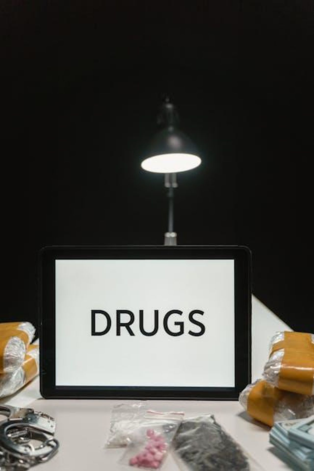 Fermate_cinque_persone_zIva1azuyj - Confidenze24 Source: MART PRODUCTION - pexels - A display of drugs, handcuffs, and contraband under a lamp in a dark room.