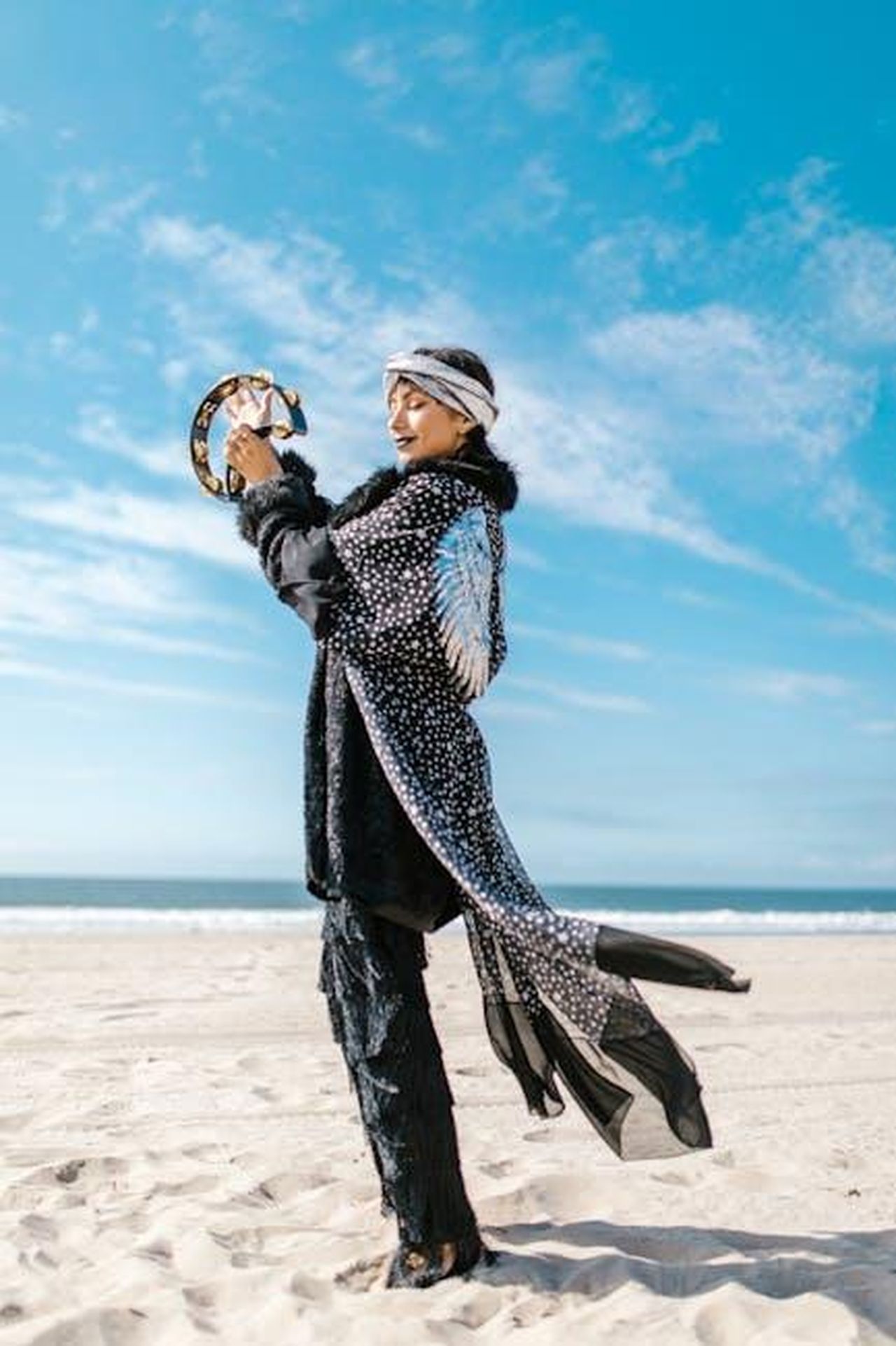 Gemelli_opportunit_in_amore_ddpcR1xDWB - Confidenze24 Source: RDNE Stock project - pexels - A woman in a patterned coat plays a tambourine on a sunny beach, embracing a spiritual vibe.
