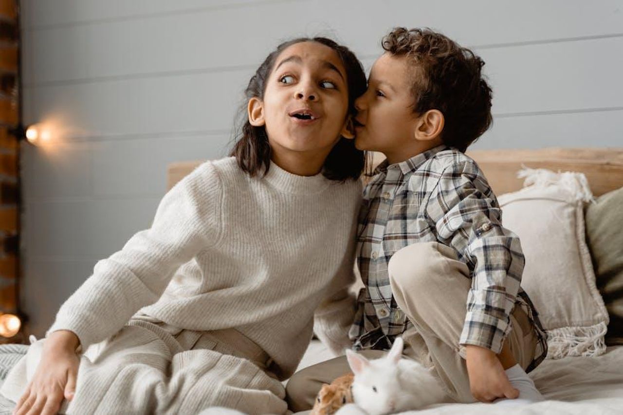Grande_Fratello_una_sorpresa_per_Helena_il_videomessaggio_della_sorella_Mariana_3Qlisd9TH8 - Confidenze24 Source: Tima Miroshnichenko - pexels - Two children whispering on a cozy bed, expressing surprise and joy in a warm, indoor setting.