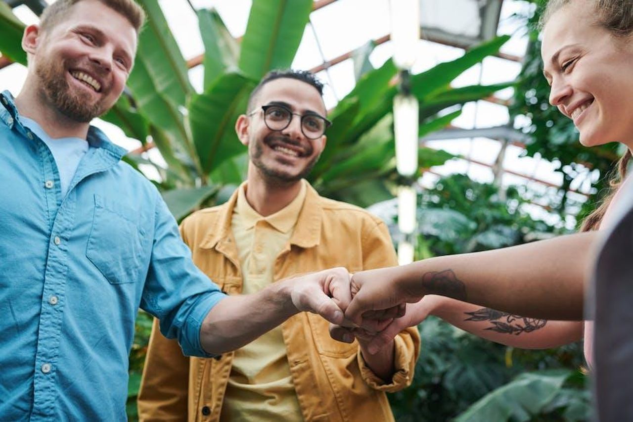 I_fan_le_inviano_affetto_UcycRJNKvE - Confidenze24 Source: fauxels - pexels - Three friends sharing a joyful fist bump in a lush indoor greenhouse environment.