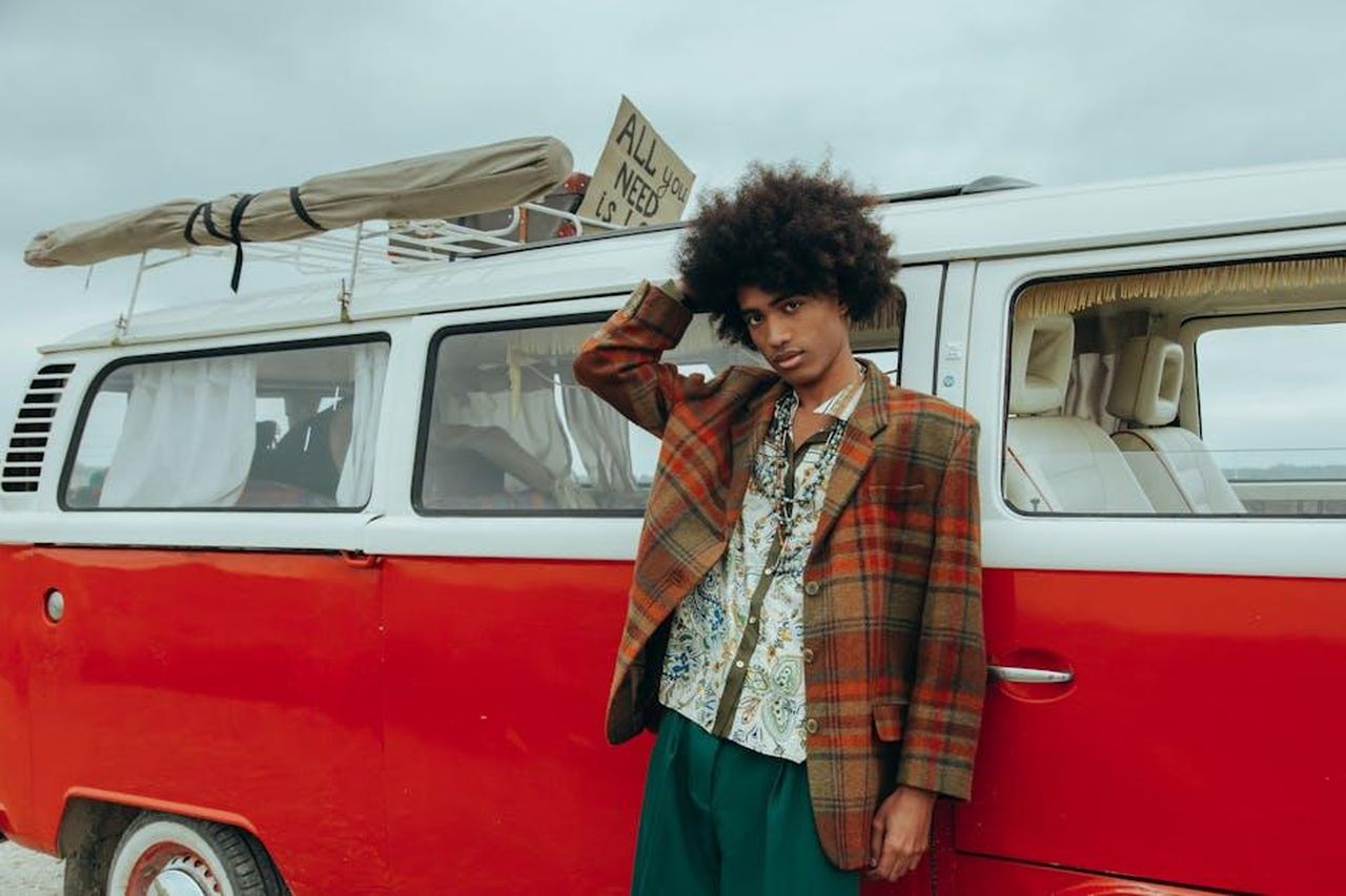 Il_Contesto_del_Tour_e_le_Polemiche_T3KxxAptHT - Confidenze24 Source: cottonbro studio - pexels - Man with afro in retro attire leaning on a vintage red van, embodying 60s hippie vibes.