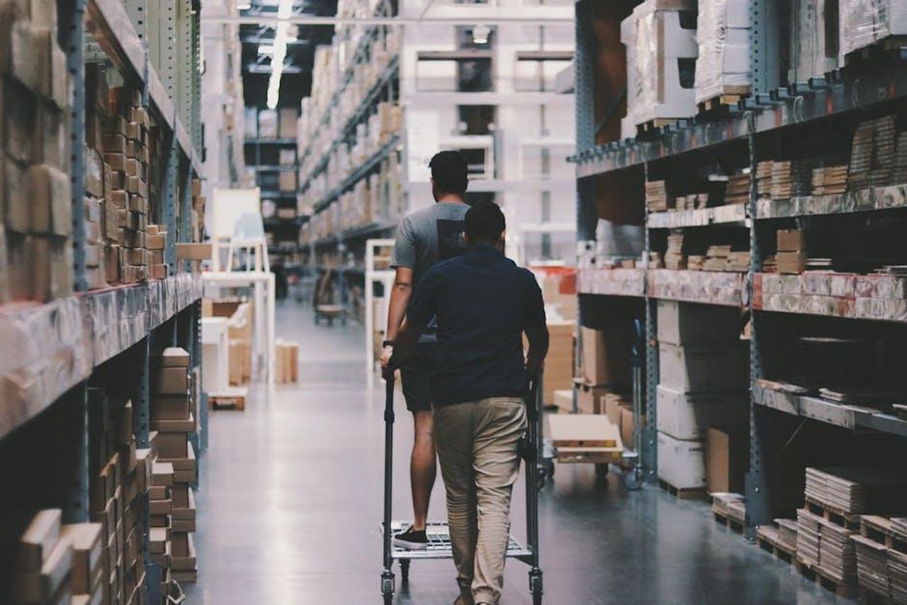 Il_Magazzino_dei_Premi_mXPO8Myq3g - Confidenze24 Source: Alexander Isreb - pexels - Two men maneuver a trolley in a large warehouse filled with boxes and shelves.
