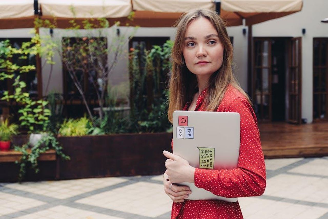Il_Paradiso_delle_signore_si_ferma_cambio_programmazione_Rai_estate_Xlr0Tpz9fv - Confidenze24 Source: Anna Shvets - pexels - A young woman in a red dress holding a laptop outdoors, looking thoughtful.
