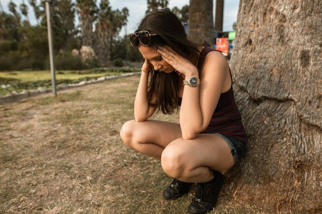 Il_cambiamento_nella_figlia_GkUoS2Wzc7 - Confidenze24 Source: RDNE Stock project - pexels - A woman kneels by a tree holding her head, expressing stress or anxiety in an outdoor park.
