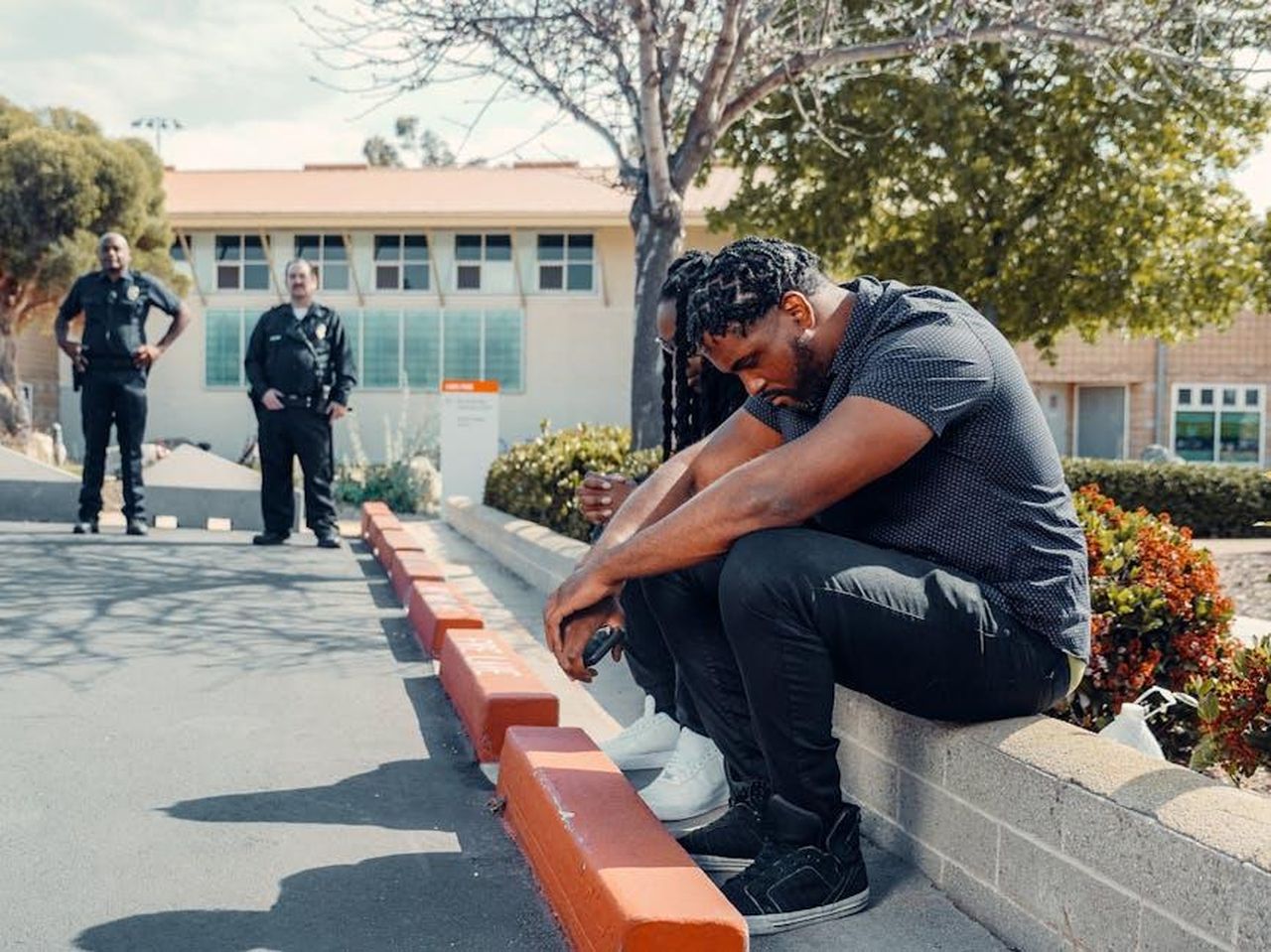 Il_primo_incontro_tra_Vick_Hope_e_Calvin_Harris_0Uvfj0FXxp - Confidenze24 Source: Kindel Media - pexels - Police officers observing two adults sitting on a curb, possibly detained, in a sunny outdoor setting.