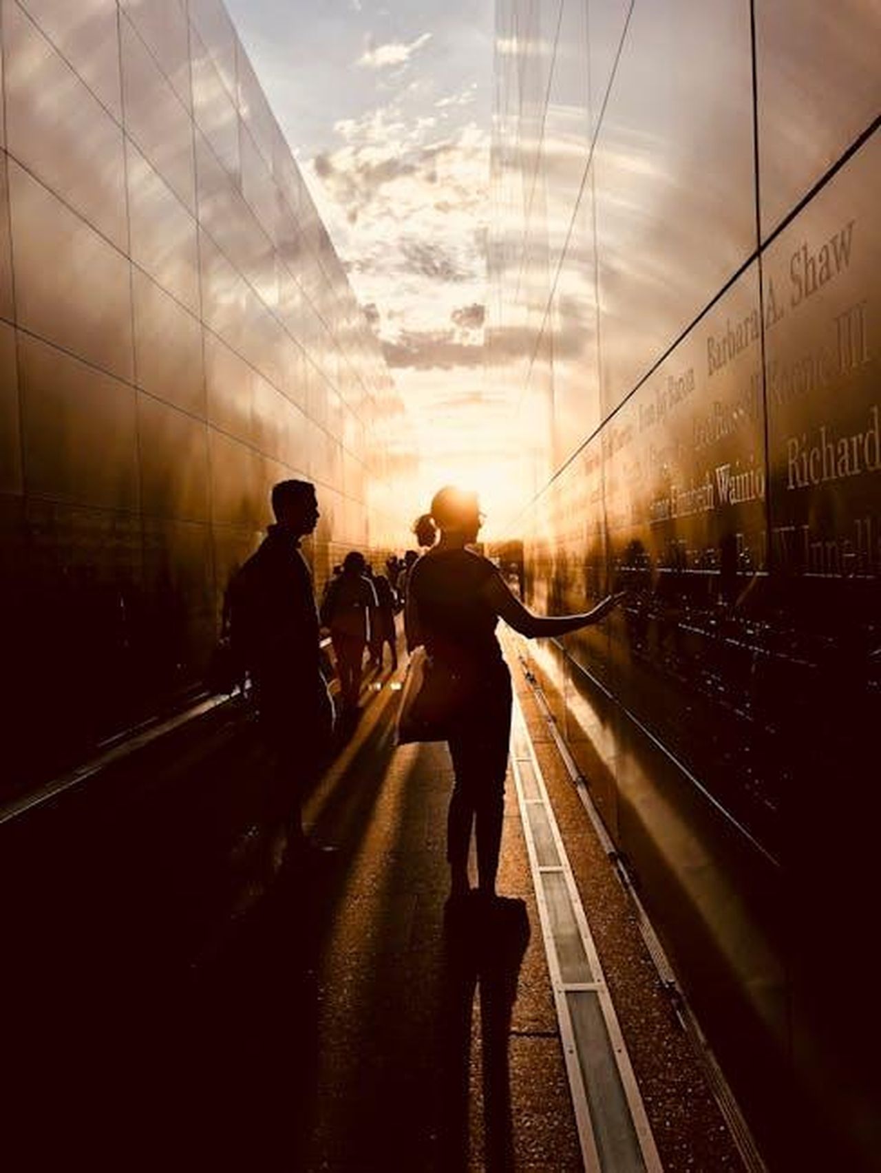 Il_ricordo_di_amici_e_colleghi_NBanXUyn3K - Confidenze24 Source: Tobe Roberts - pexels - Silhouettes of people at a reflective memorial wall during sunset, creating a contemplative atmosphere.