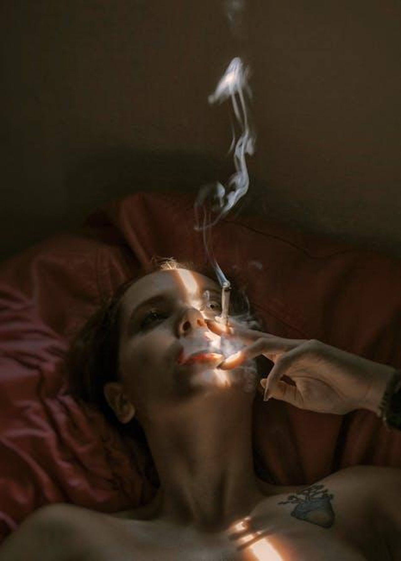 Il_rimorso_che_non_lascia_in_pace_Sezai_j9pMRV6djx - Confidenze24 Source: Bianca Salgado - pexels - A woman lying on a bed indoors, smoking a cigarette with dramatic lighting casting shadows on her face.
