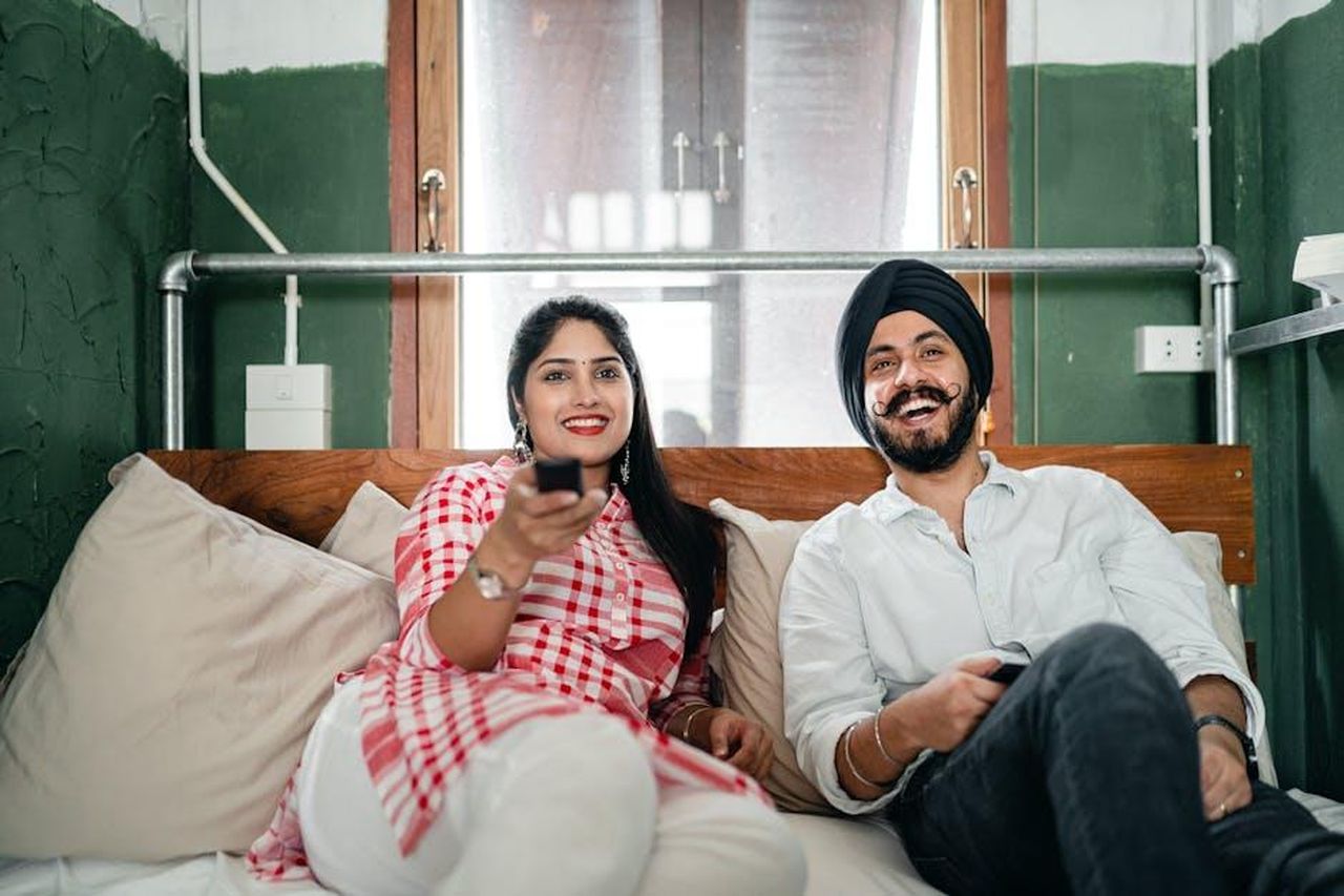 Il_successo_della_soap_in_tv_e_streaming_online_FZP0vfwR1z - Confidenze24 Source: Ketut Subiyanto - pexels - Cheerful wife with bindi on forehead wearing plaid tunic with white trousers using TV remote control for channel switching while lying on bed with laughing Sikh husband in turban with stylish beard and twisted mustache