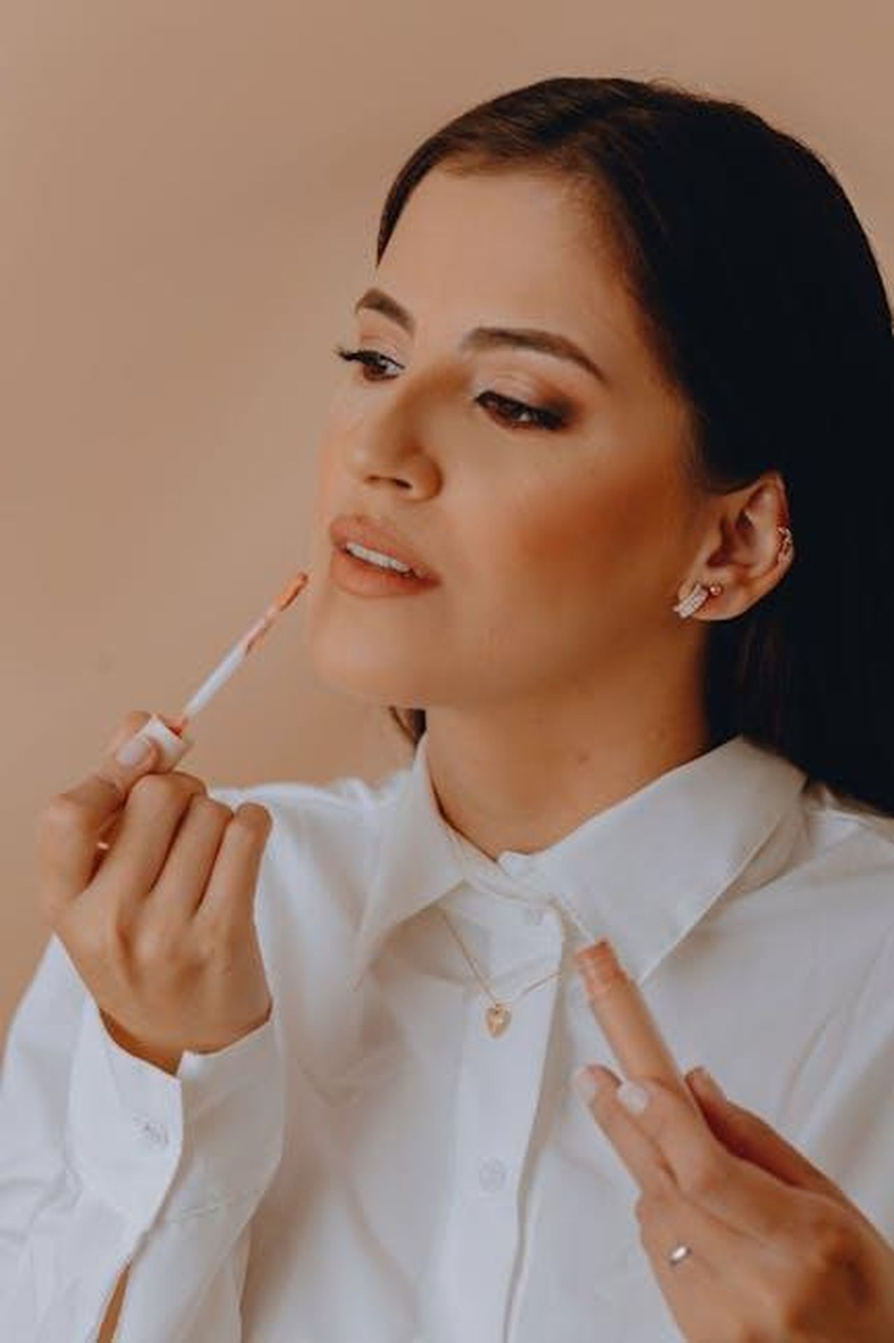 Il_talento_di_Olympia_nel_trucco_JDcHQ0Pym2 - Confidenze24 Source: Adriele Nenes - pexels - A woman in a white shirt gracefully applies lip gloss against a soft beige background.