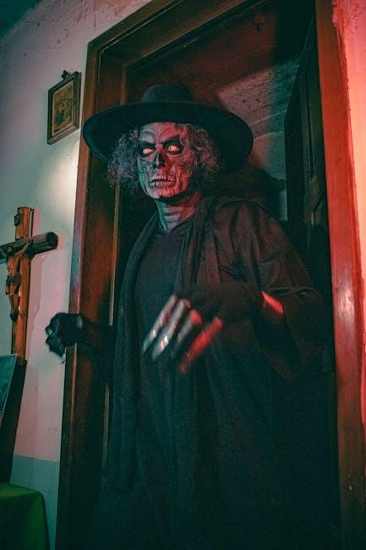 Il_timore_di_Javier_ACbZlmqAU6 - Confidenze24 Source: Moisés Fonseca - pexels - A sinister figure in skull makeup and a hat emerges from a dimly lit doorway, creating a chilling atmosphere.