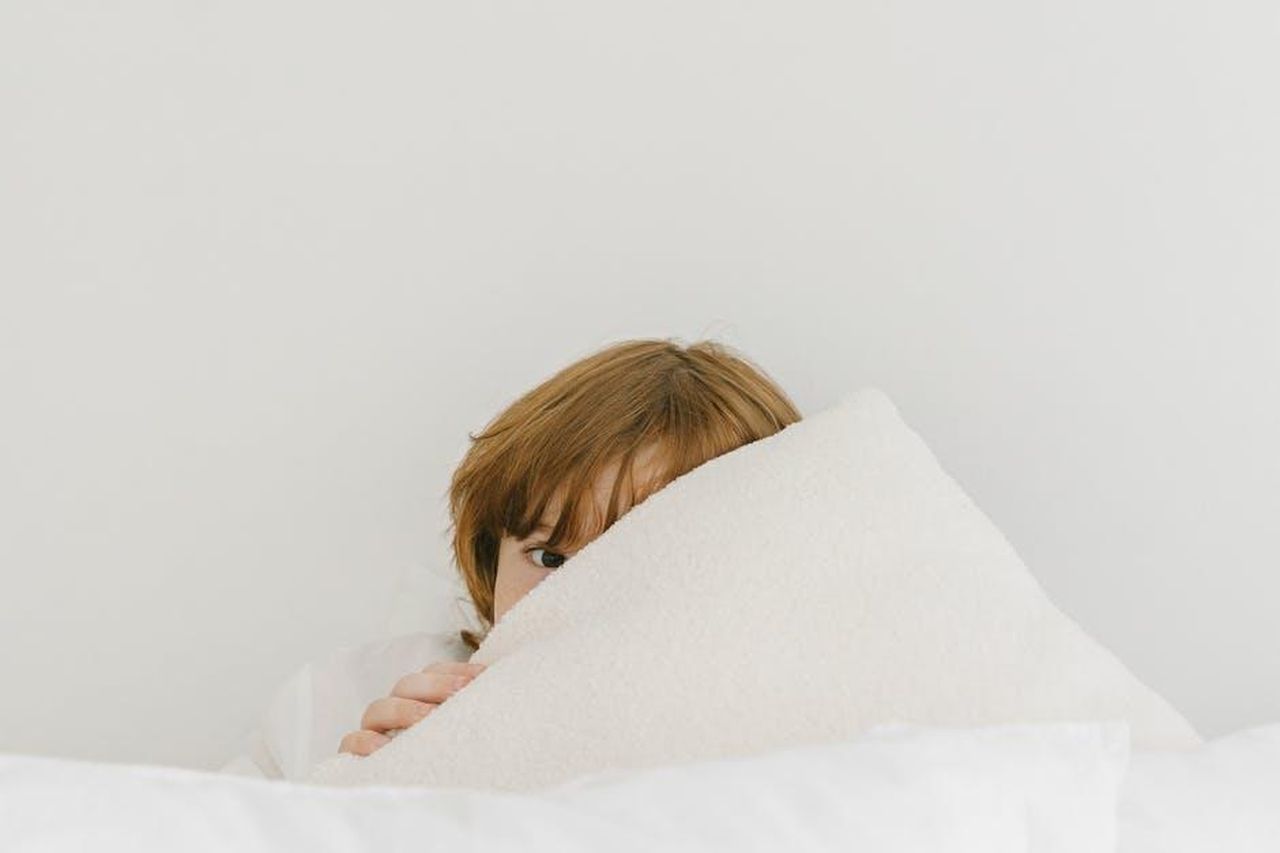 Jana_Romulo_e_Alonso_proteggeranno_Pia_KKnbrQ6bl5 - Confidenze24 Source: Mikhail Nilov - pexels - A shy child peeks over a soft white pillow in a cozy indoor setting.