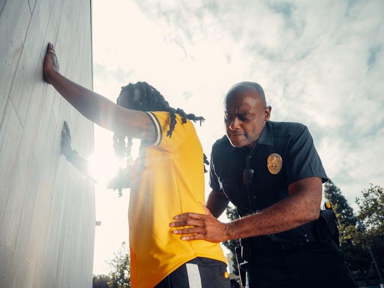 La_chiamata_alla_polizia_e_il_passato_oscuro_di_Tom_g4HtV0z61j - Confidenze24 Source: Kindel Media - pexels - A police officer conducts a frisk search on a suspect wearing a yellow shirt outdoors.