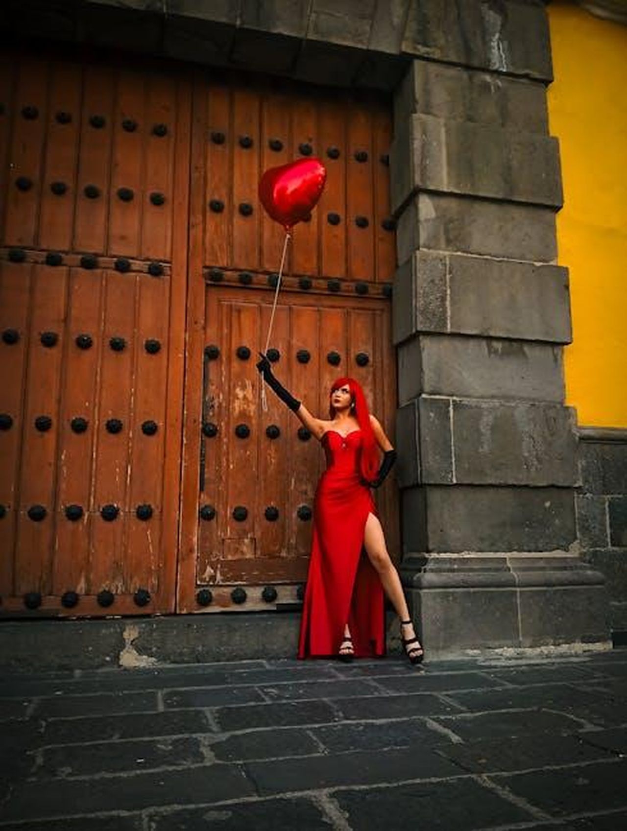 La_nuova_relazione_della_star_di_Call_The_Midwife_Helen_George_vvtn67ijBc - Confidenze24 Source: Carlos Jesús Alonso Aguilar - pexels - Stylish woman in red dress poses with a heart balloon against a historic wooden door.