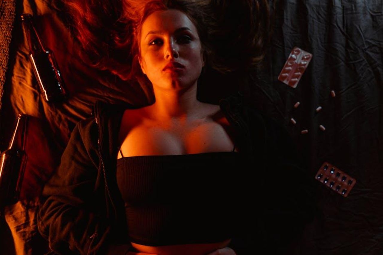 La_reazione_inaspettata_del_marito_parole_che_feriscono_uoRVCmdXWz - Confidenze24 Source: MART PRODUCTION - pexels - A woman lying on a bed surrounded by bottles and pills, illuminated by moody lighting.