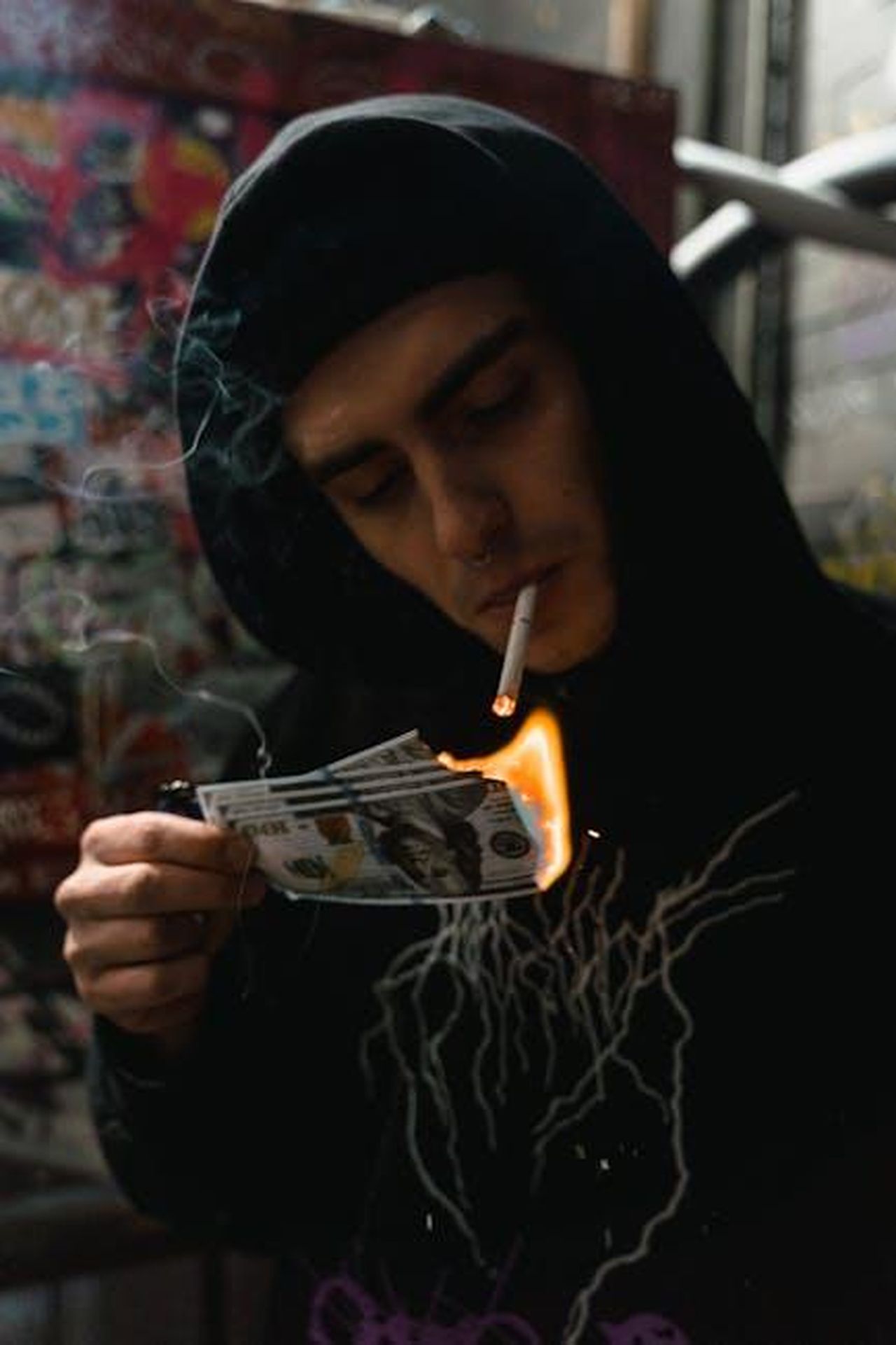 Le_conseguenze_del_tradimento_zI8zYAZ0b2 - Confidenze24 Source: MART PRODUCTION - pexels - A man in a black hoodie burns money with a cigarette, showcasing rebellion.