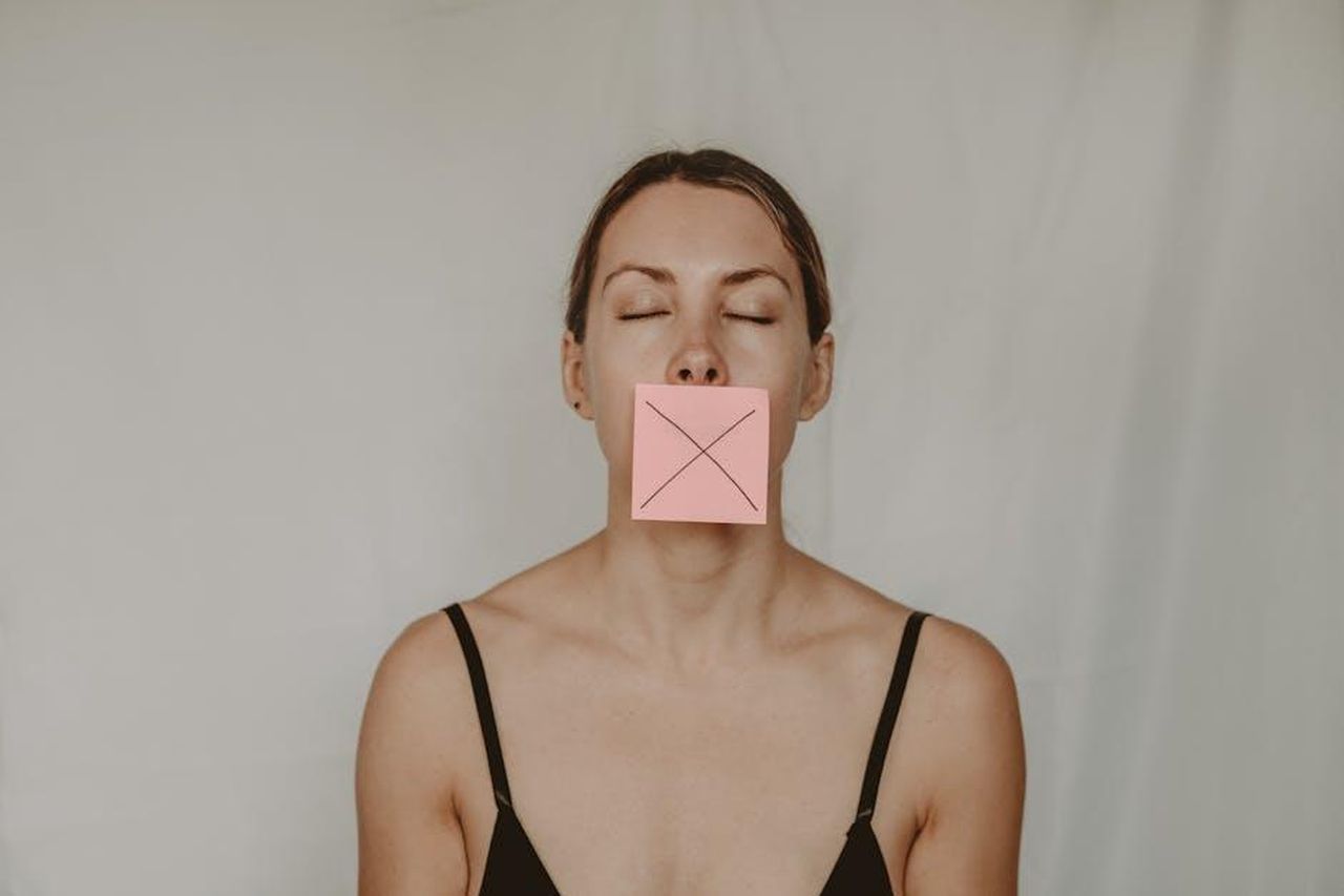 Le_parole_toccanti_del_conduttore_gsdoGO5PkP - Confidenze24 Source: Anete Lusina - pexels - Young slender woman with closed eyes and mouth covered with sticky note showing cross on white background