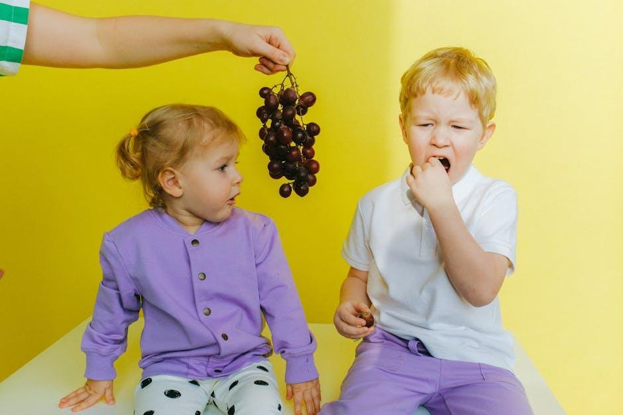 Manuel_geloso_di_Pino_cDHban26iV - Confidenze24 Source: Anna Shvets - pexels - A boy enjoying grapes while sitting next to his curious sister indoors against a yellow background.