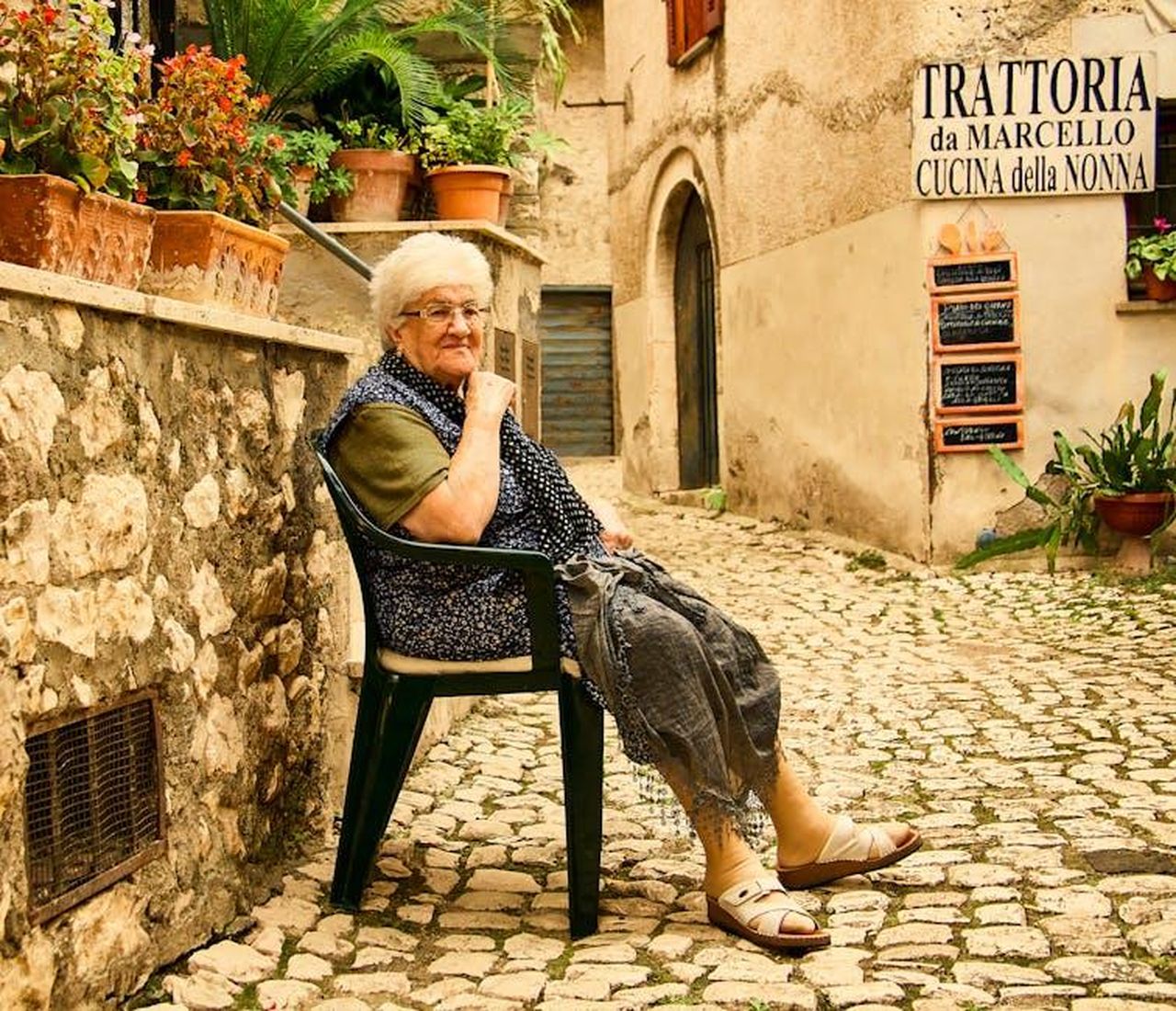Marcello_riceve_un_riconoscimento_ma_non_vuole_festeggiarlo_la_trama_del_28_febbraio_5stVzCfh2e - Confidenze24 Source: Wycher van Vliet - pexels - Senior woman in Italian street, sitting by trattoria, surrounded by potted plants.