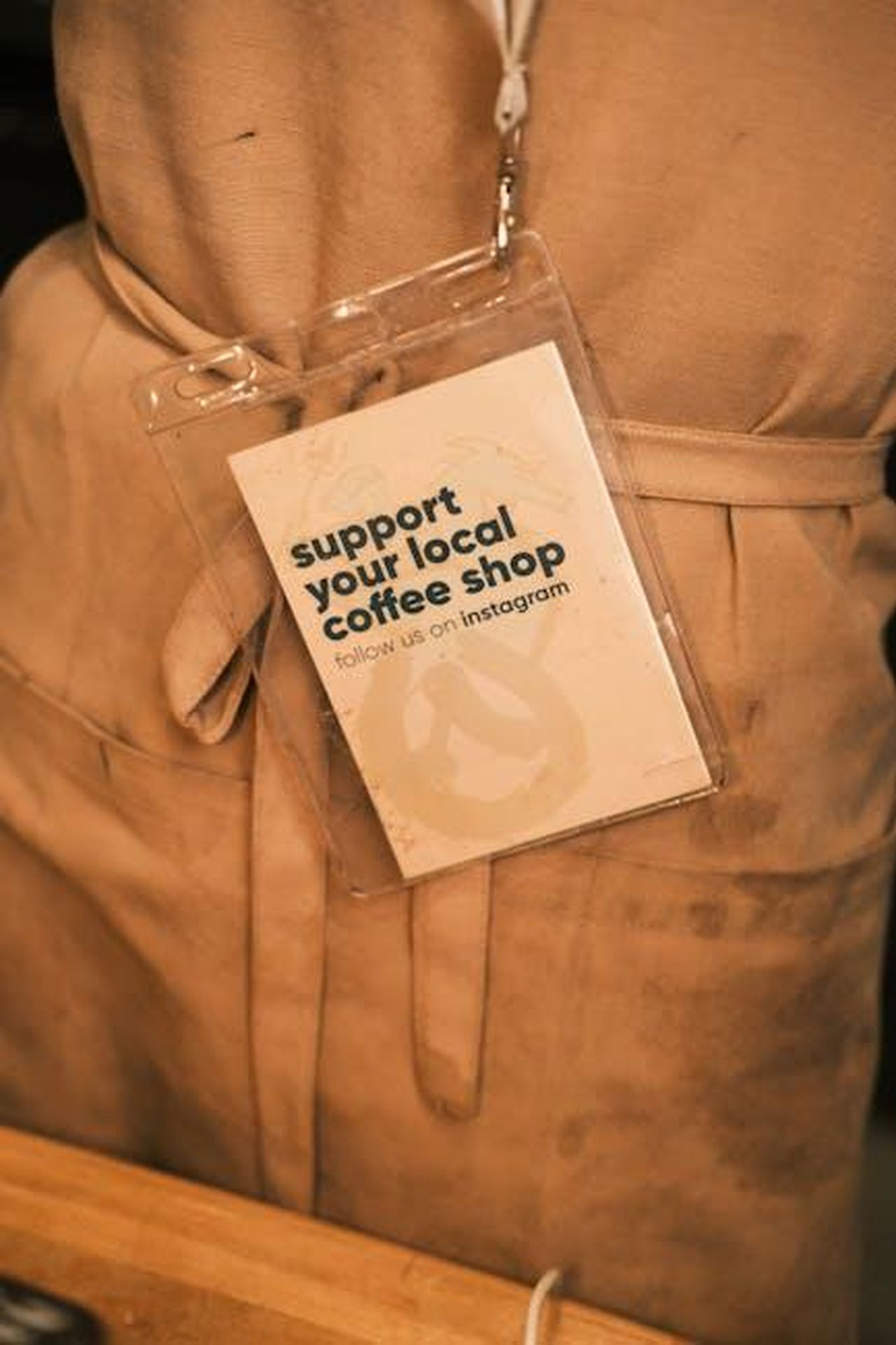 Meghan_e_il_suo_supporto_tramite_Instagram_nPj2NDSpgn - Confidenze24 Source: Cemrecan Yurtman - pexels - Close-up of a barista apron with a tag promoting local coffee shop support.