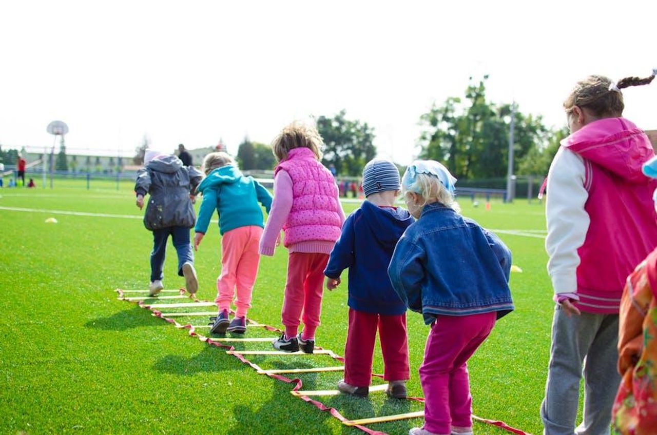 Momenti_speciali_con_i_bambini_O5yDEzcfpi - Confidenze24 Source: Lukas - pexels - Children enjoy an outdoor activity on a grassy field, stepping over a ladder.