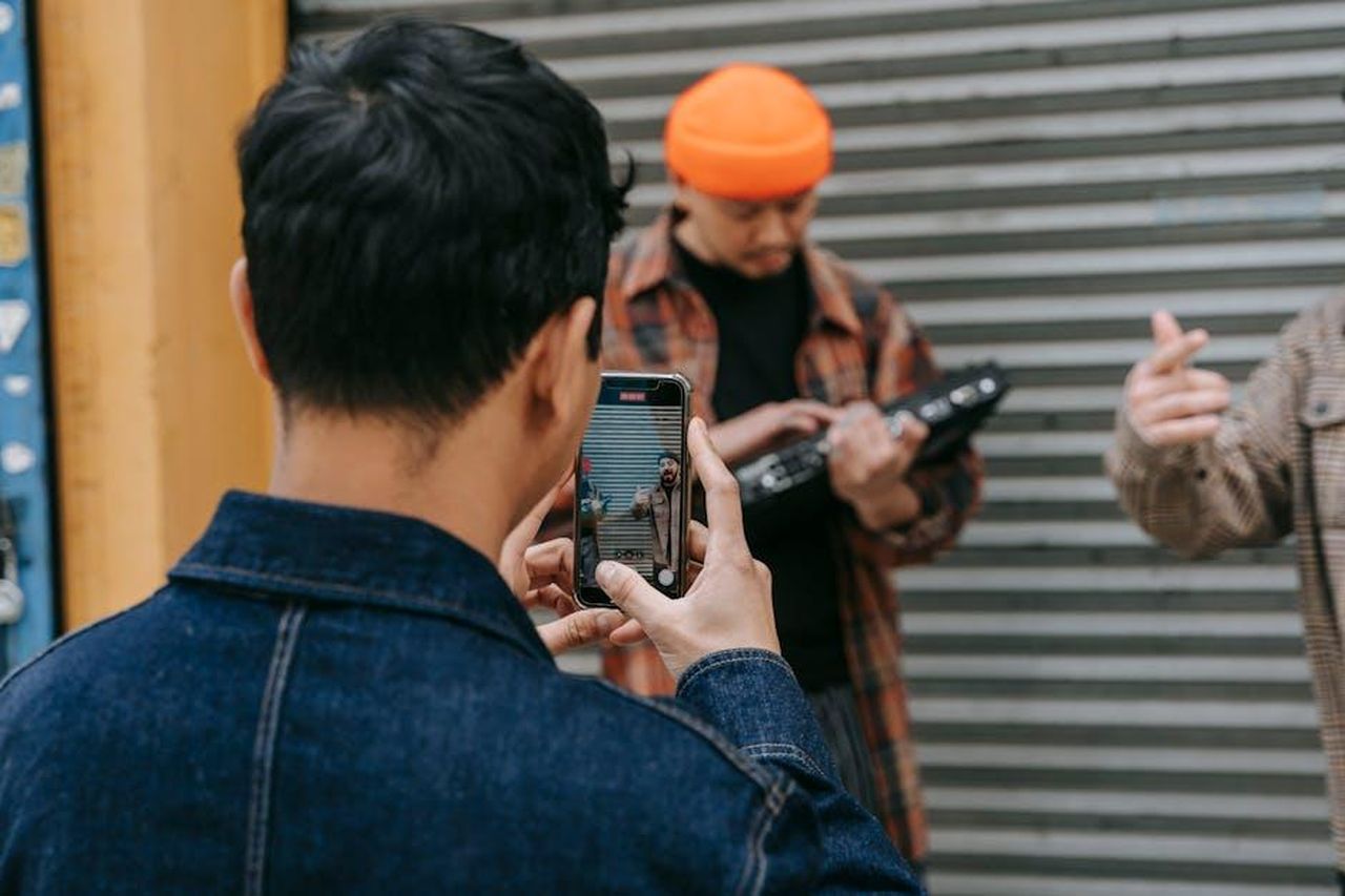 Momenti_virali_della_serata_oR5JMWTktQ - Confidenze24 Source: Anna Pou - pexels - A person takes a photo of street musicians using a smartphone in an urban setting.