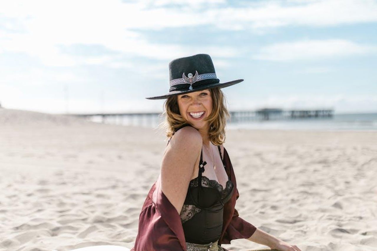 Oroscopo_degli_ultimi_quattro_segni_ZQIJsIac3A - Confidenze24 Source: RDNE Stock project - pexels - A woman smiles joyfully on a sunny beach wearing a stylish hat and a casual outfit.