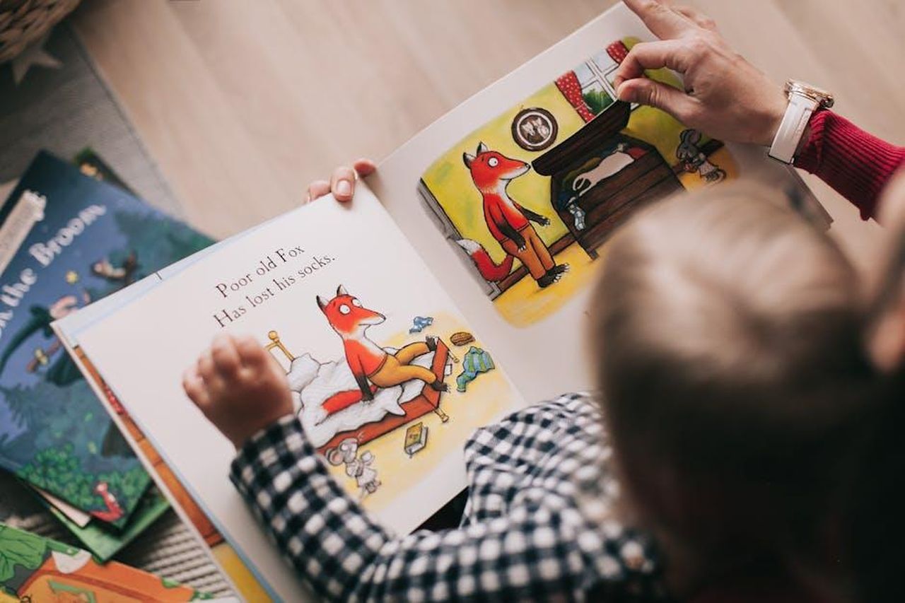 Oyku_viene_affidata_a_Selime_PdjgHYqta1 - Confidenze24 Source: Lina Kivaka - pexels - A child and adult reading an illustrated storybook together in a cozy setting.