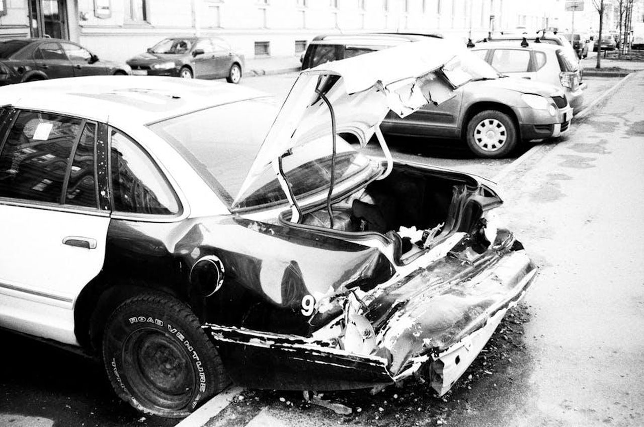 Oylum_difesa_da_Behram_dopo_aver_rischiato_di_essere_investita_da_unauto_wZUuSaqApi - Confidenze24 Source: Aleksandr Neplokhov - pexels - A black and white photo of a wrecked car on an urban street, highlighting vehicle damage.