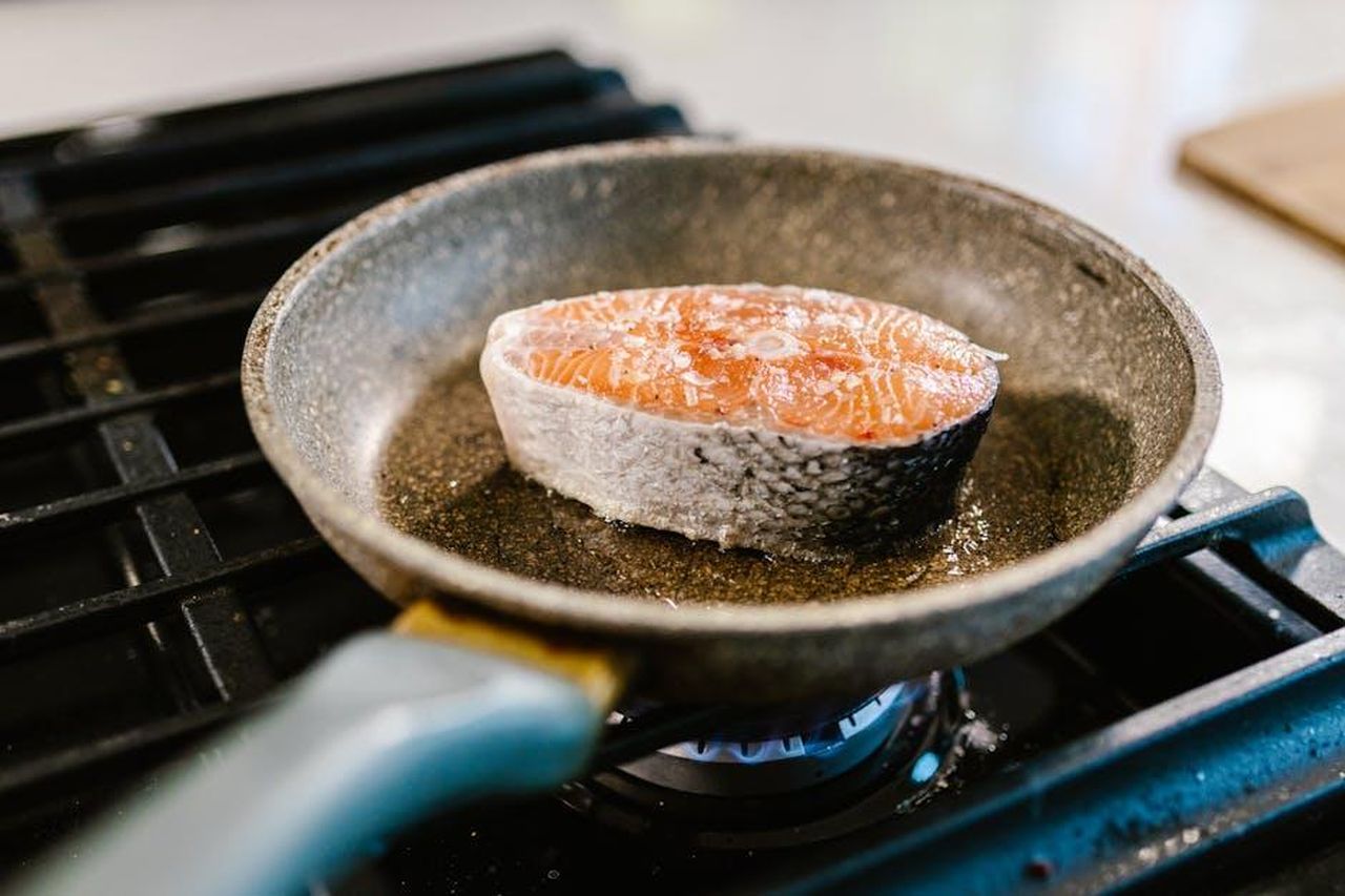 Ricetta_Salmone_arrosto_lento_con_riso_verde_labneh_capperi_e_nasturzi_ifNlbDeRbq - Confidenze24 Source: RDNE Stock project - pexels - A salmon steak is searing in an iron frying pan on a stove, showcasing cooking detail.
