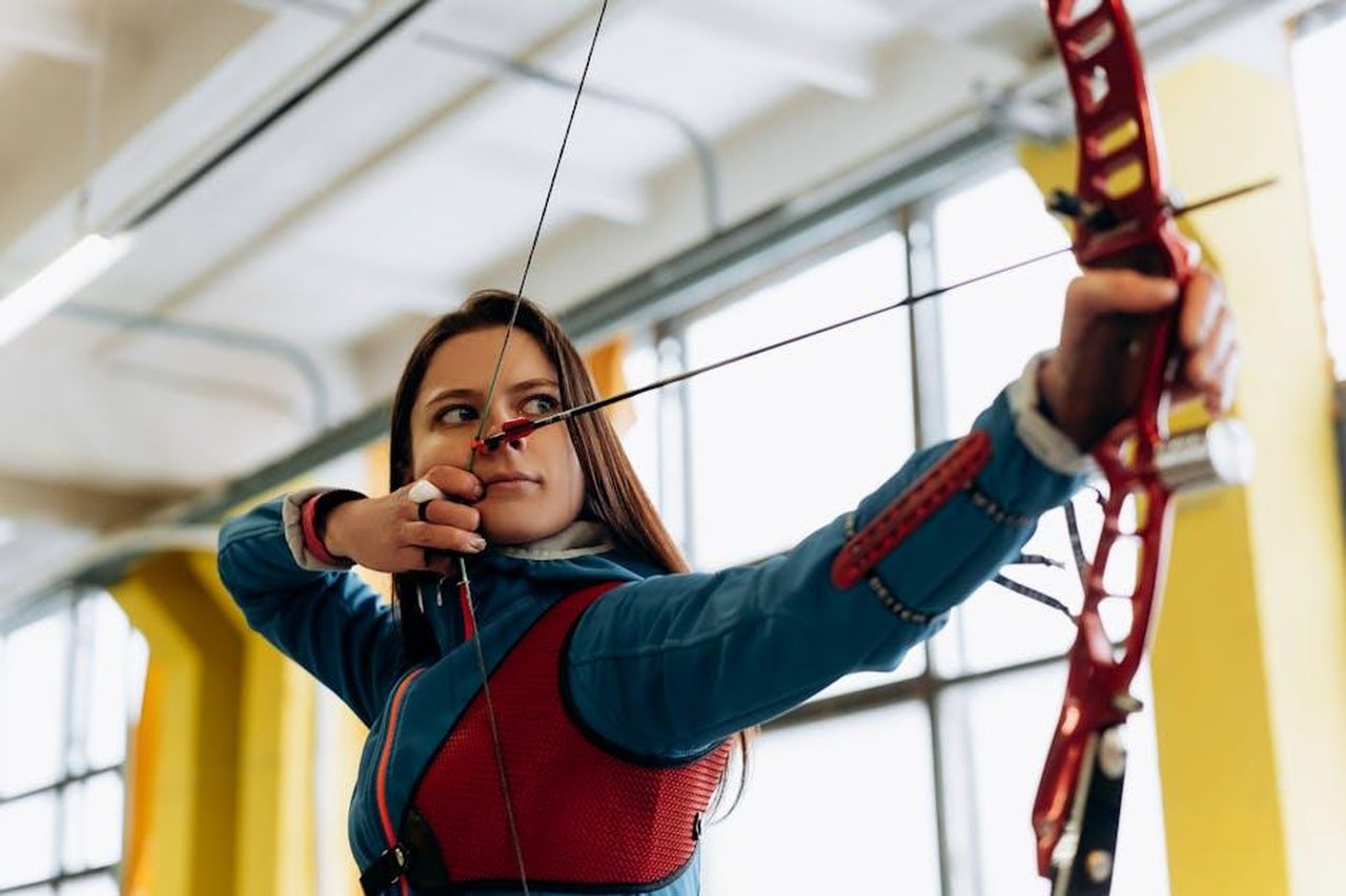 Rocco_Hunt_con_Clementino__Yes_I_know_my_way_Pino_Daniele_x3kjosYMep - Confidenze24 Source: Mikhail Nilov - pexels - Young female archer in blue aiming a bow and arrow indoors, showcasing concentration and athleticism.