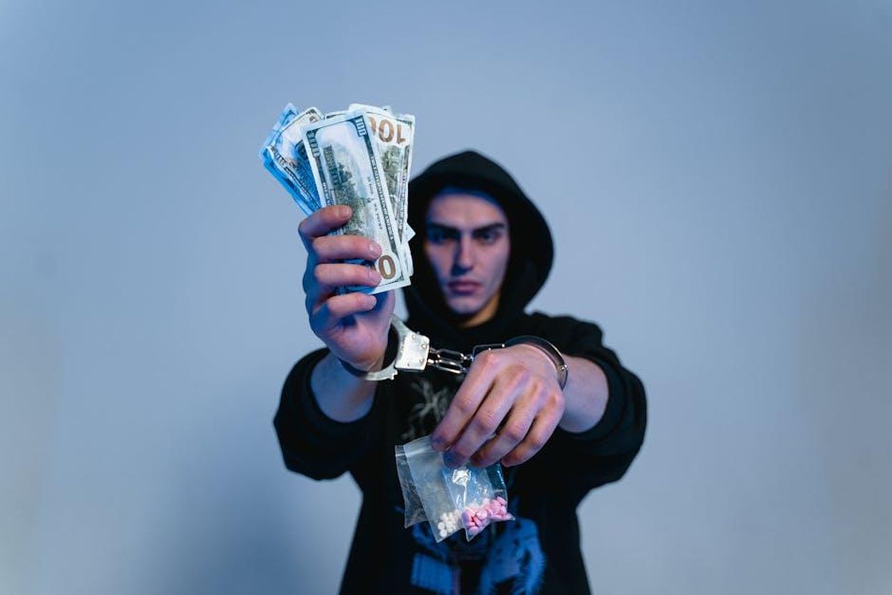 Ruolo_di_Rita_nel_traffico_di_cocaina_ICwyFOF3dg - Confidenze24 Source: MART PRODUCTION - pexels - Handcuffed man in a hoodie holding cash and drugs, depicting crime and addiction.
