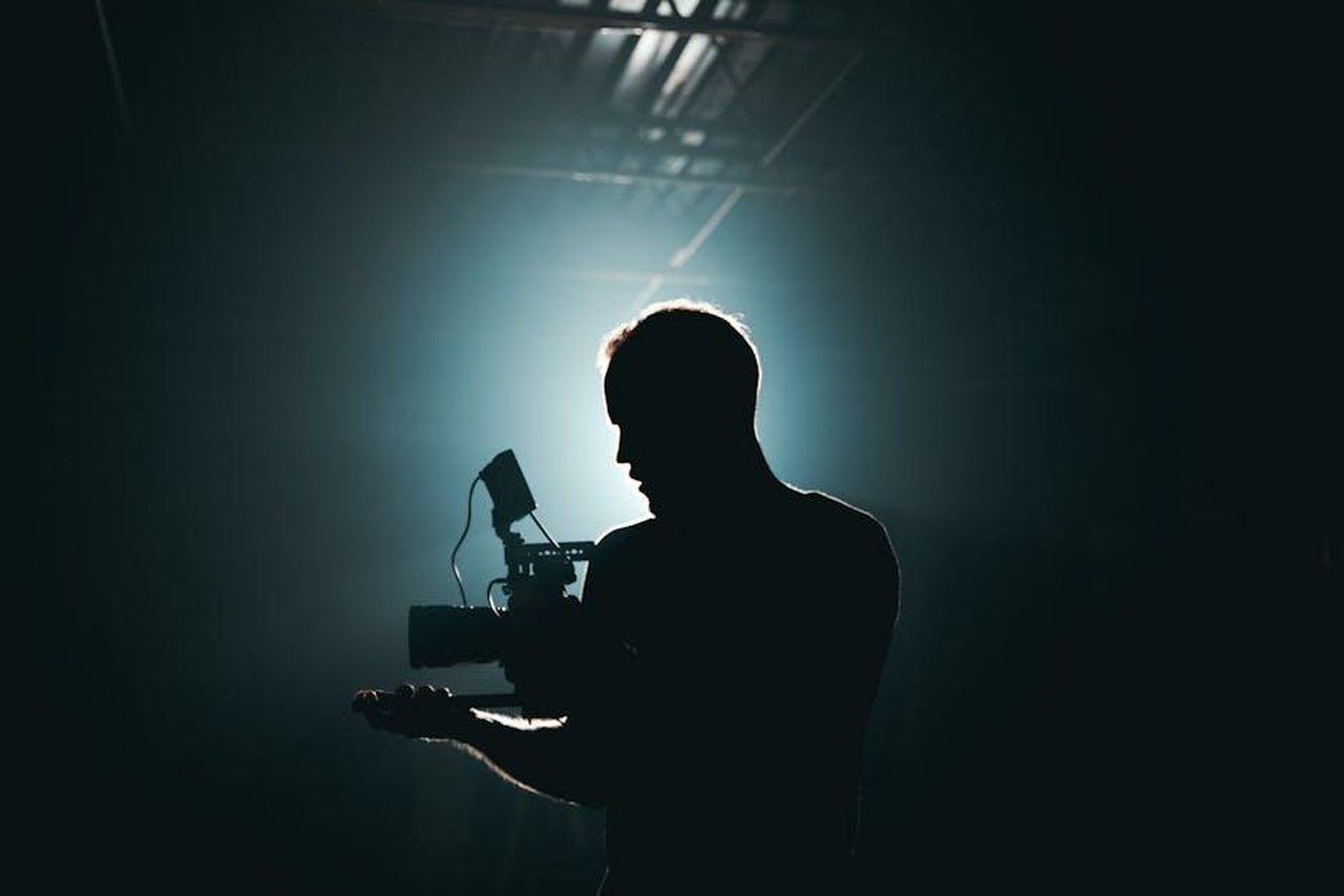 Settembre_2015_Premiere_di_Dateline_aJ46vXUx2F - Confidenze24 Source: Kyle Loftus - pexels - Silhouette of a cinematographer with camera gear in a dramatic, low light setting.