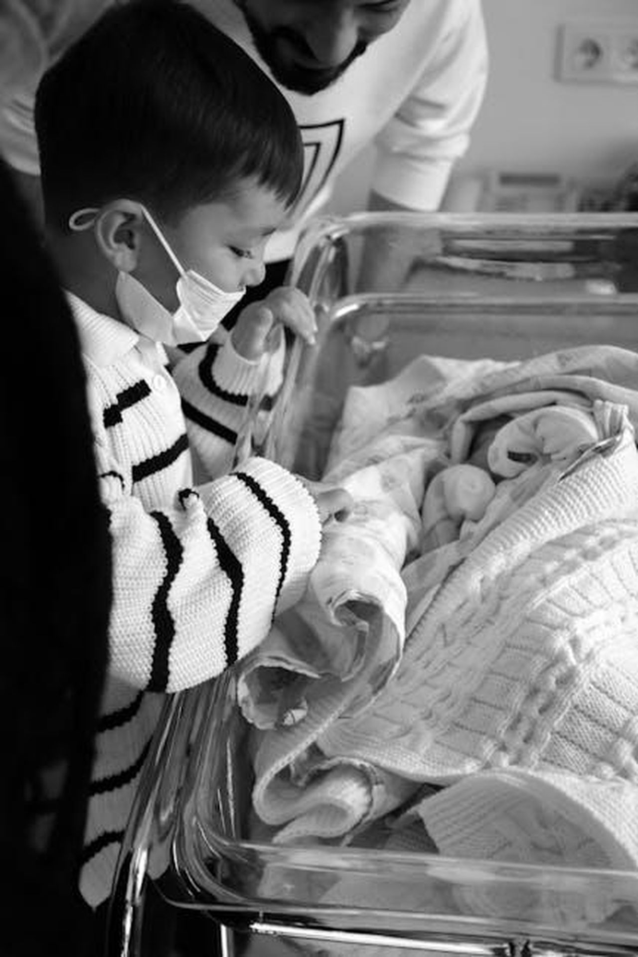 Tarik_scopre_di_Ozan_in_ospedale_grazie_a_Nazan_V1F6eC7cKh - Confidenze24 Source: Dilara Doğar - pexels - A young child wearing a mask looking at a newborn baby in a hospital setting. Black and white image.