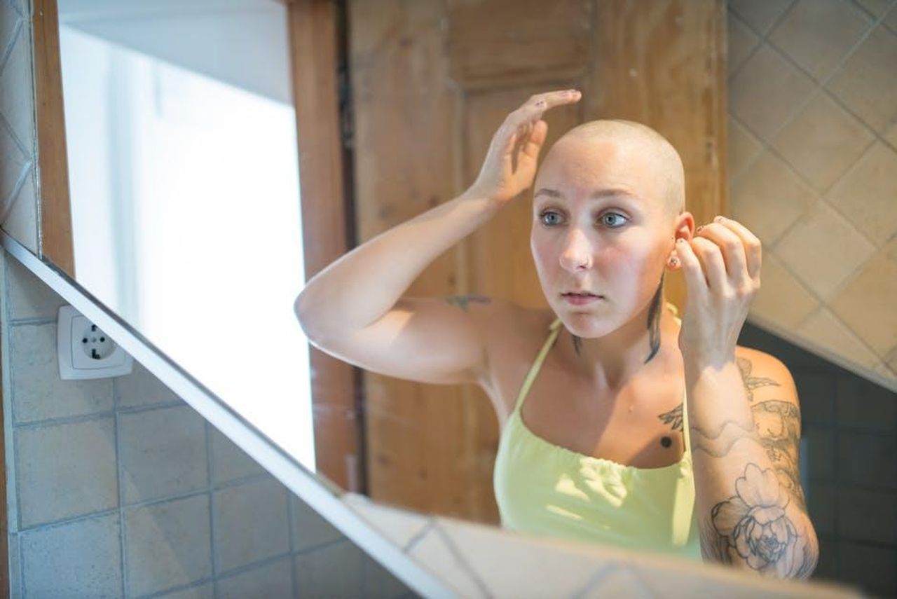 Tatuaggi_piercing_e_fede_un_conflitto_tra_tradizione_e_modernit_vlQVpCWoyn - Confidenze24 Source: Kampus Production - pexels - A tattooed woman with a shaved head adjusts her appearance in a mirror.