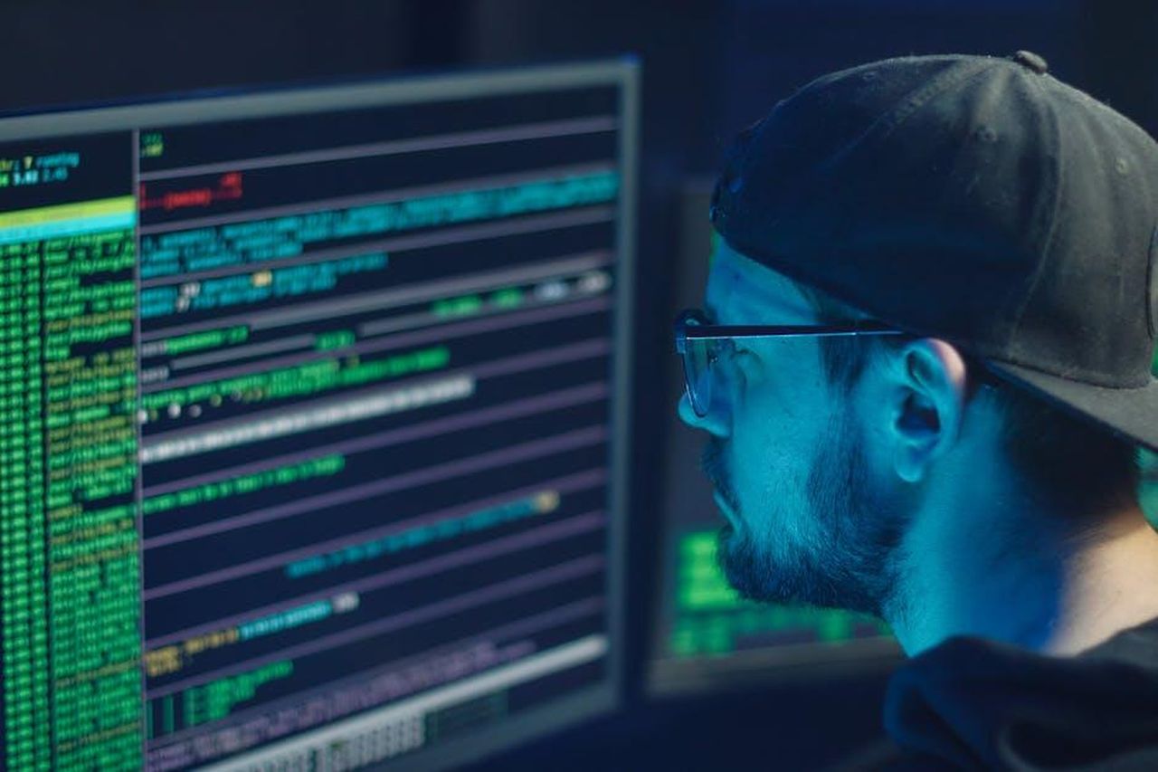 Tolga_subisce_un_attacco_hacker_AezZ5WYamG - Confidenze24 Source: Mikhail Nilov - pexels - A cybersecurity expert inspecting lines of code on multiple monitors in a dimly lit office.