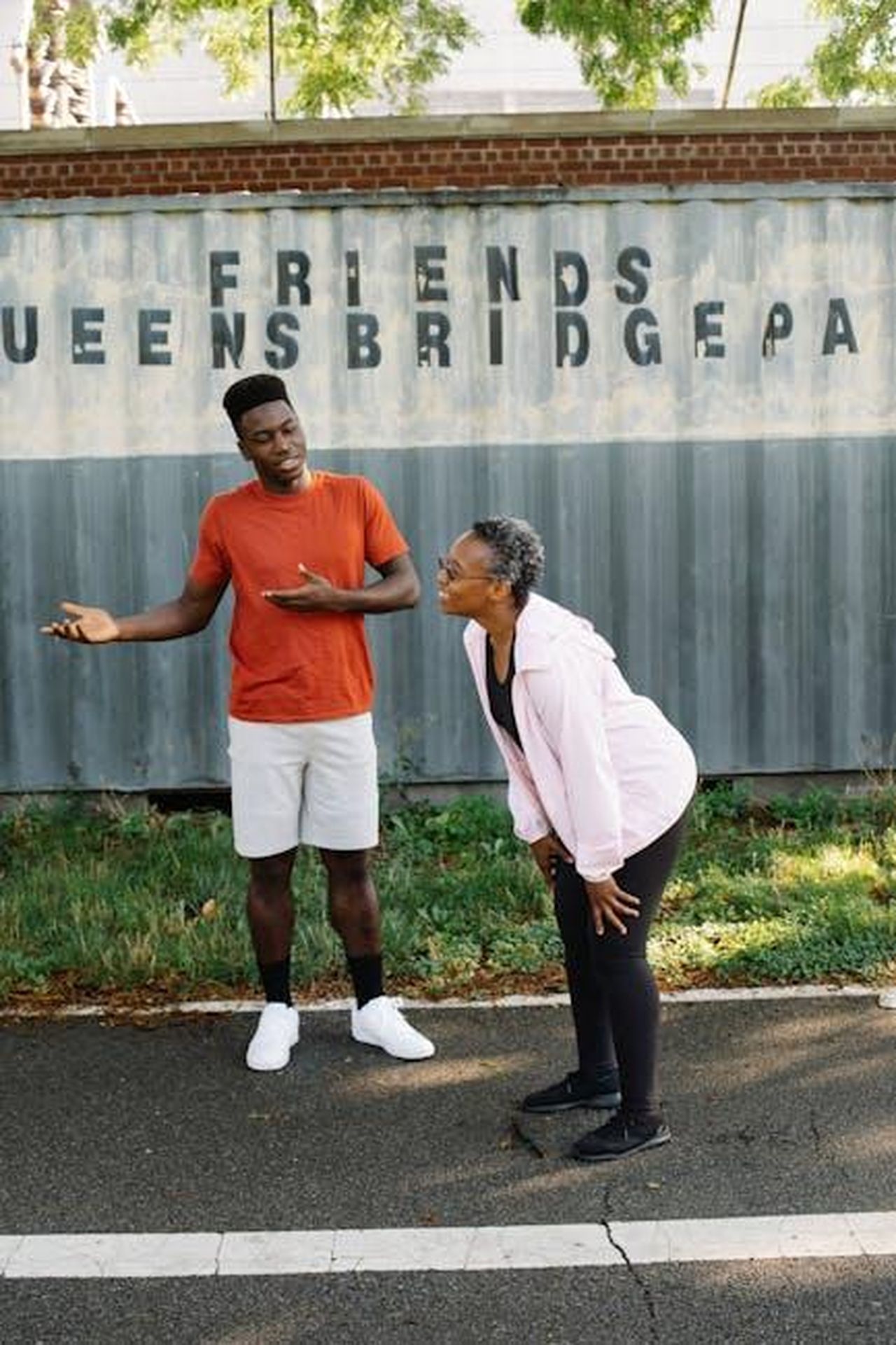 Ultimo_saluto_nel_quartiere_di_Queens_f5ue3lULtj - Confidenze24 Source: Liliana Drew - pexels - Two friends share a lively conversation outdoors by the Queensbridge Park sign.