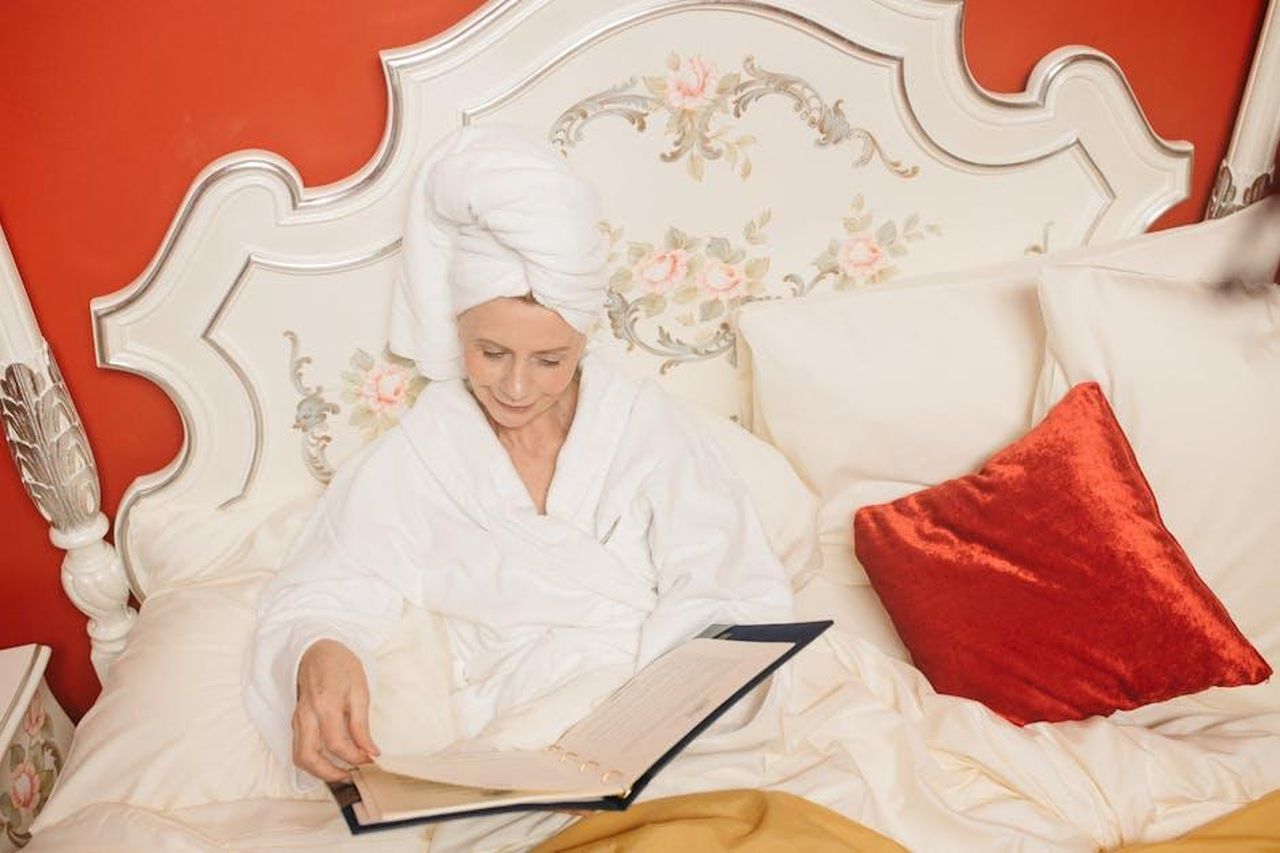 15_agosto_2023_un_altro_incontro_tra_Brady_e_Shayk_RzXqaHWNrD - Confidenze24 Source: Anastasia Shuraeva - pexels - A woman in a white robe relaxes in a beautifully decorated bedroom, reading a menu.