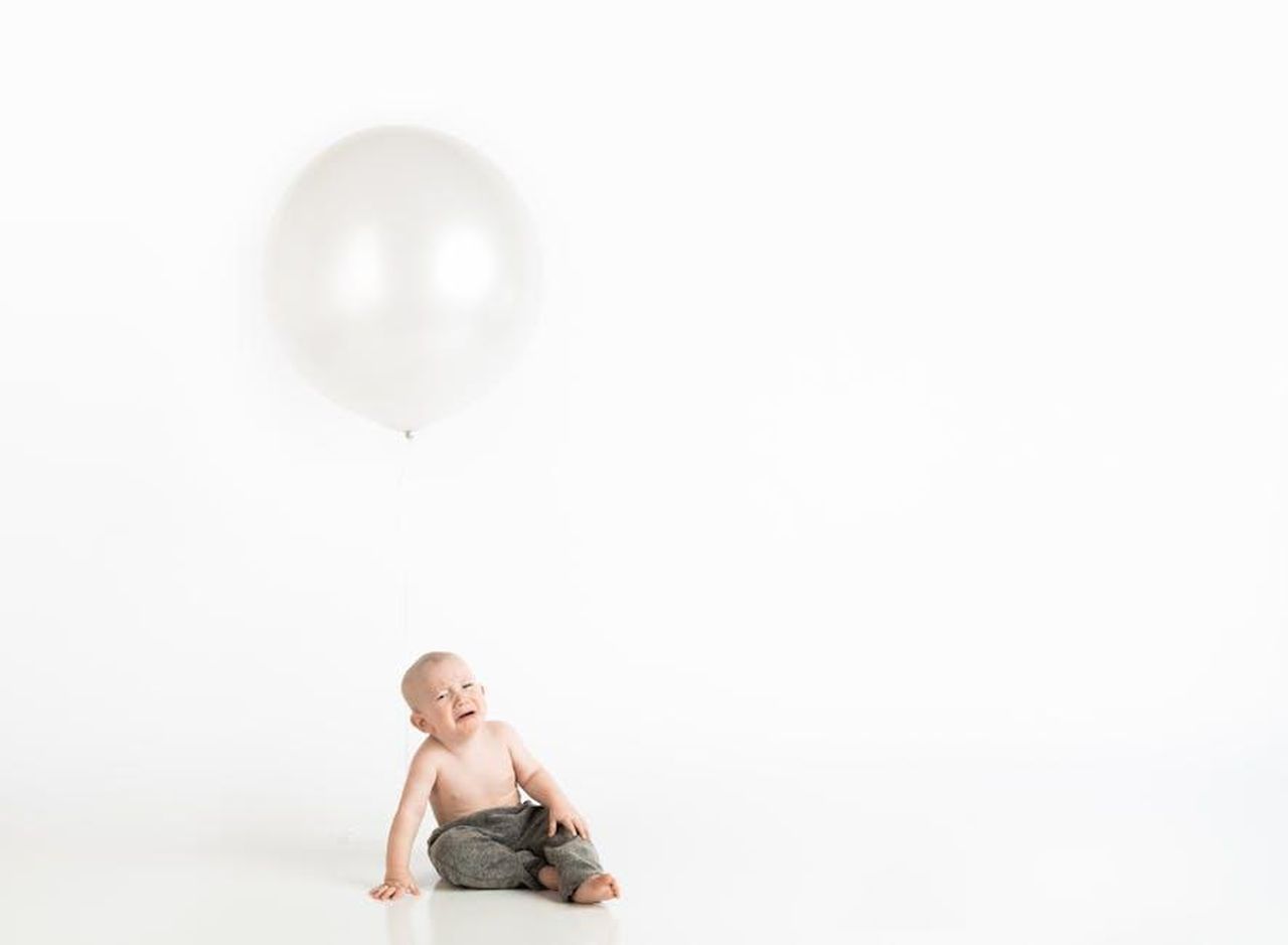 2_Georgia_parla_di_genitorialit_da_sola_LgyDBzVyPn - Confidenze24 Source: Henley Design Studio - pexels - A baby sitting on the floor crying next to a large white balloon in a minimalist studio setting.